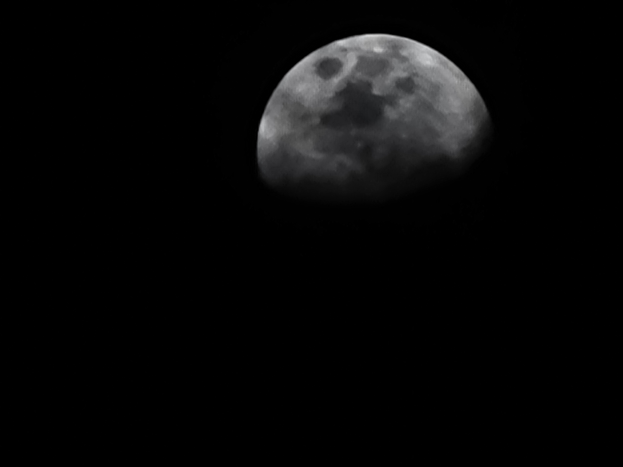 The image is a photograph of the moon against a black sky. The moon is partially illuminated, showing craters and dark patches on its surface. The lighting suggests it is either a crescent or gibbous phase. The image is cropped, with the moon occupying the right side of the frame and the left side being entirely black space. There are no discernible features of Quarteira, Portugal, or any other urban environment in the image.