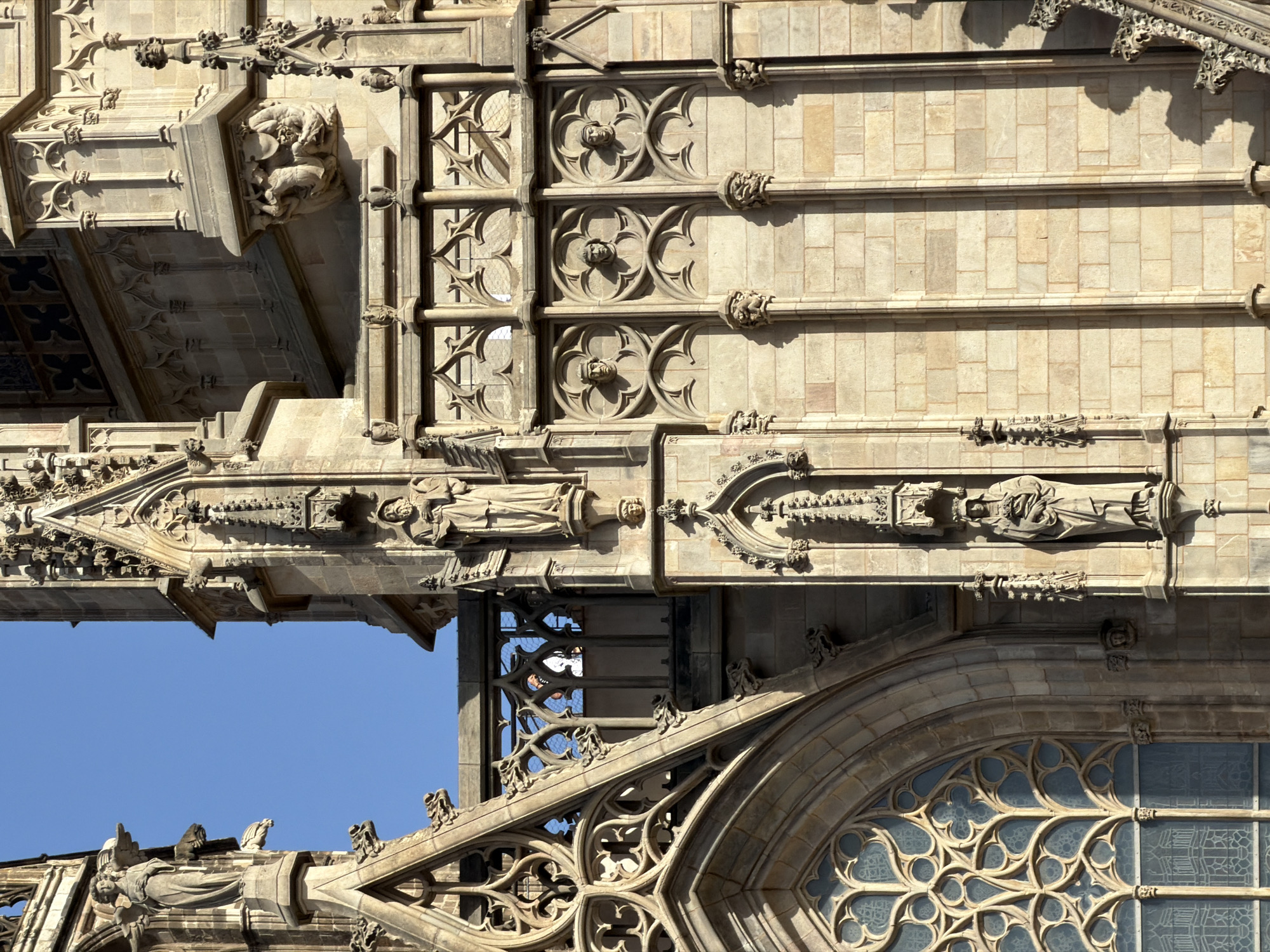 The image shows a detailed view of a Gothic cathedral facade in Barcelona, Spain, bathed in bright sunlight. The architecture is characterized by intricate stonework, pointed arches, and ornate carvings. A prominent feature is a large rose window with delicate tracery. Statues of figures, possibly saints or angels, are mounted on pedestals and niches along the facade, some appearing to hold objects. Decorative gargoyles are also visible, typical of Gothic architecture, serving as both ornamentation and water spouts. The stone blocks forming the walls are light brown and clearly visible, showcasing the construction materials. The sky above is a clear, vibrant blue. The overall impression is one of grandeur and historical significance, highlighting the architectural artistry of the cathedral.