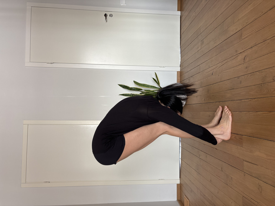 An adult female is depicted indoors, performing a forward bend exercise. She wears a long-sleeved black top and black shorts, standing barefoot on a light brown wooden plank floor. Her torso is bent forward at the waist, with her head lowered and arms extending towards her feet. Her dark hair is pulled back into a ponytail, hanging towards the floor. The background features plain white walls with white baseboards and two white doors. The door on the right is equipped with a silver-colored handle and escutcheon plate. A tall snake plant, housed in a white pot, is visible between the two doors. The indoor setting does not provide specific visual cues to identify its precise location within the city of Chaikovsky, Russia.