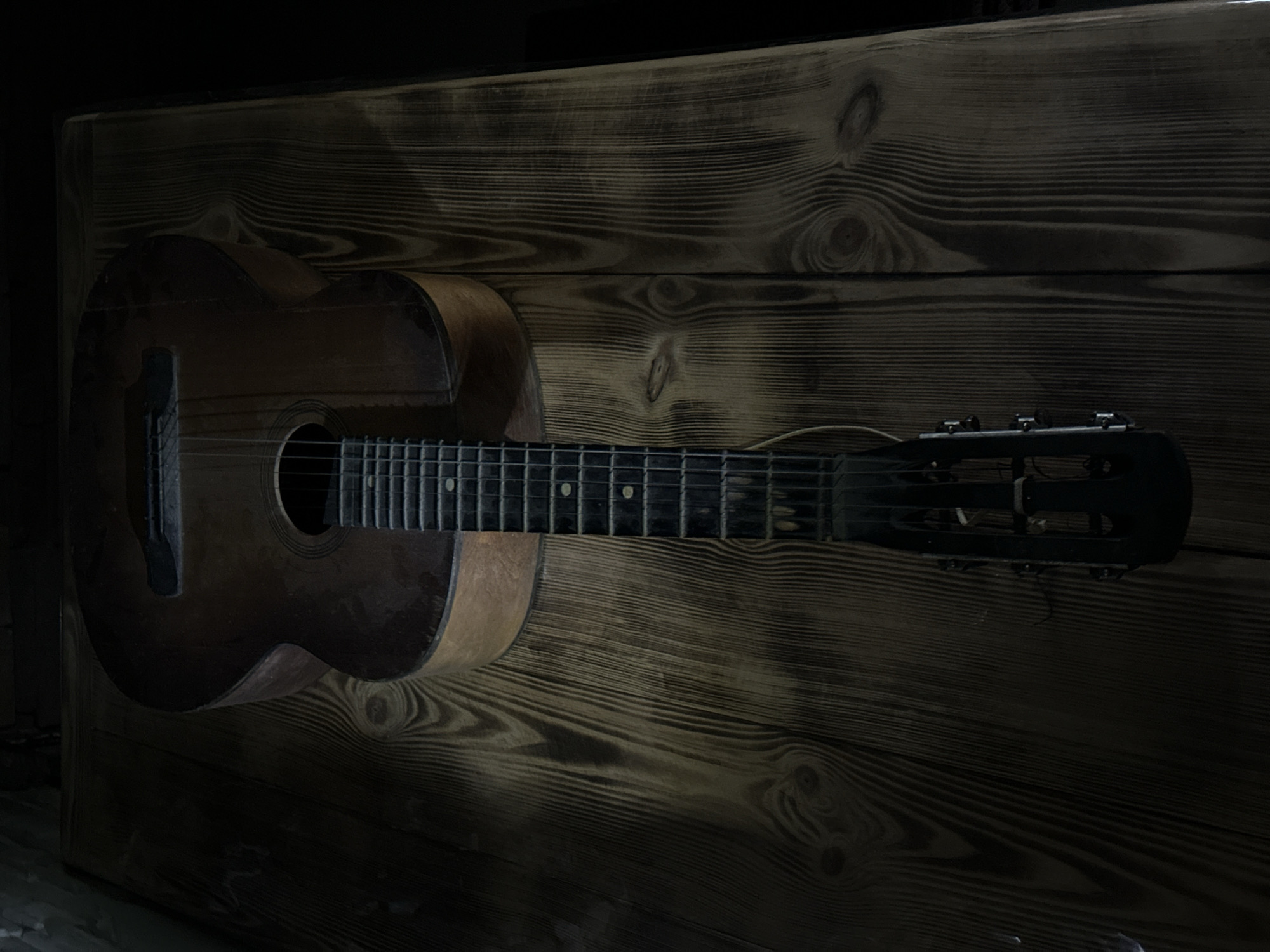 The image shows an acoustic guitar lying on a wooden table. The guitar is dark brown and appears to be old. The table is made of rustic, weathered wood with a prominent grain pattern. The lighting is dim, with a spotlight highlighting the guitar. The scene is forensic and matter-of-fact. There are no people or actions depicted in the image. The location of the image is not specified in the provided data, therefore it is not possible to provide details about the city of Luhansk, Ukraine.