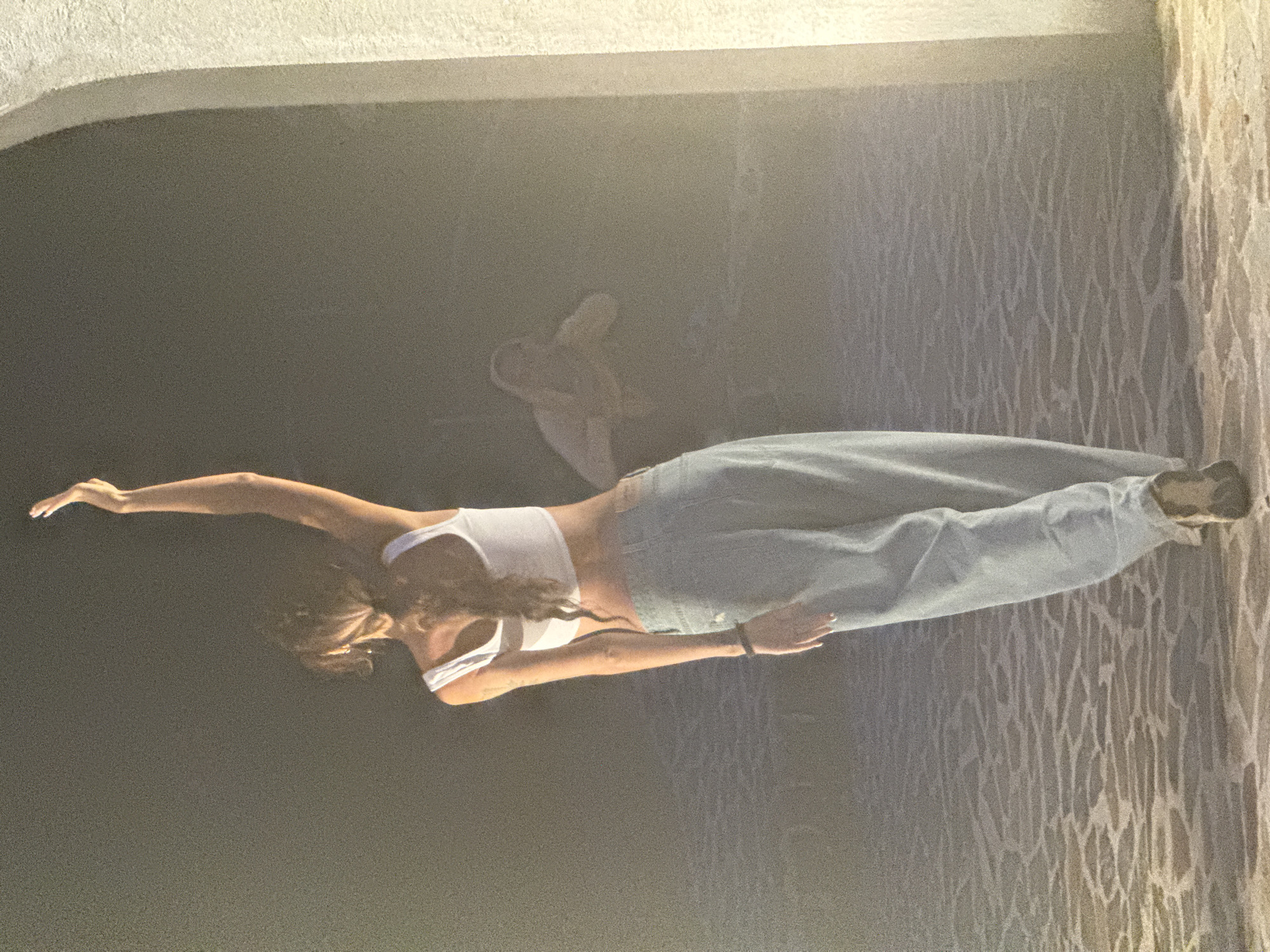 A young woman is posing for a photograph in what appears to be an outdoor architectural setting. She is wearing a white crop top and light blue, wide-legged jeans. Her right arm is extended outward, while her left arm is positioned towards her side. The background consists of a dark grey or black surface on one side and a wall with a textured pattern on the other. The setting could be a partially covered or enclosed space. In the background, it is hard to tell what is happening but it appears to show an older version of the girl sitting in the background. There is no explicit evidence to confirm the exact location as Binh Lu, Vietnam, or other related details without additional information.