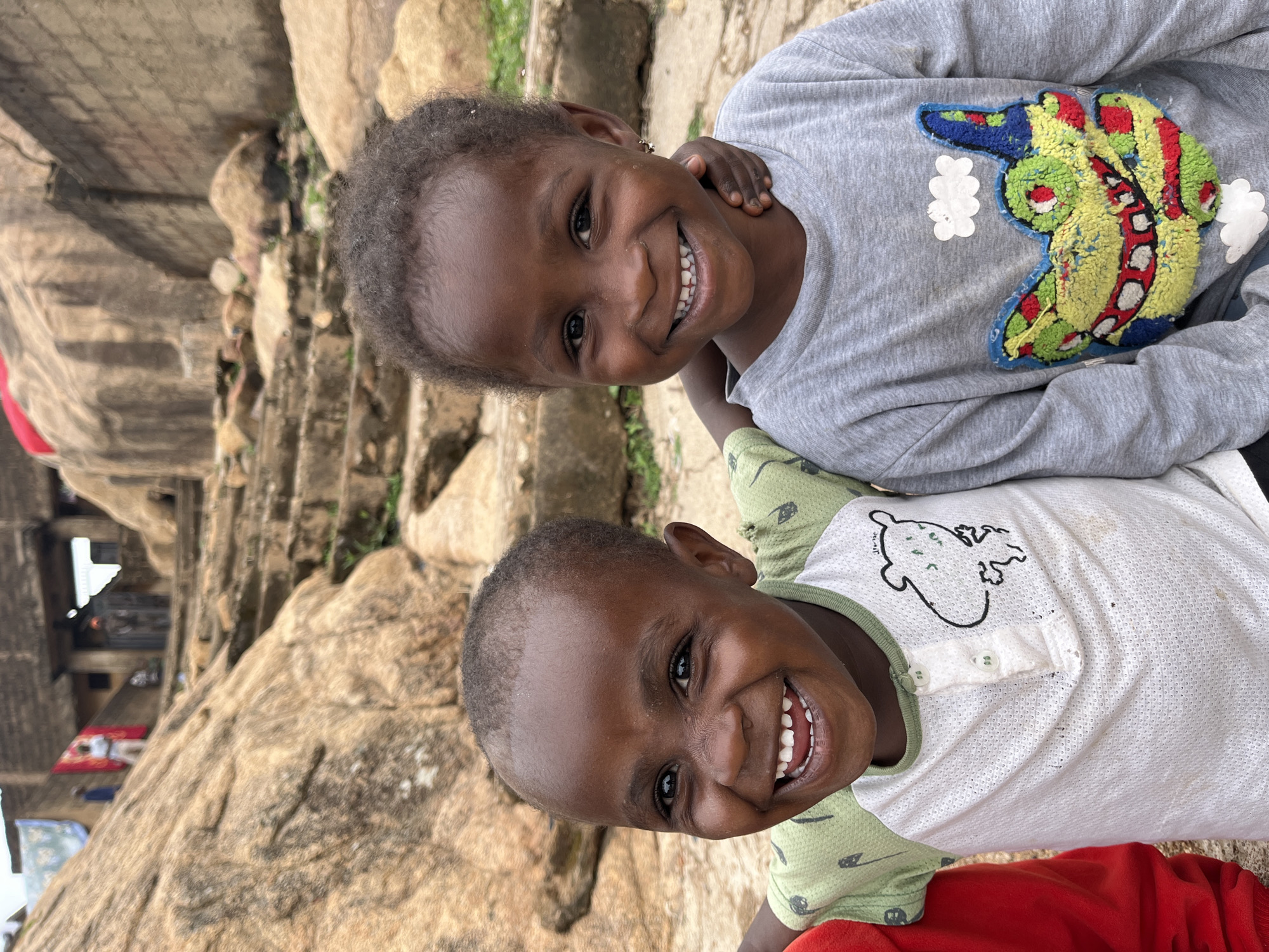 Two smiling young children, likely siblings, pose closely together outdoors in Gura Top, Nigeria. The foreground reveals a rocky, mountainous terrain, while an edifice is seen on the left side of the frame. The child on top is wearing a gray long-sleeved shirt featuring a colorful graphic. The child below wears a white, buttoned shirt with a green patterned collar and sleeves. Both children have dark skin and short, dark hair. The expressions on their faces suggest happiness and warmth.