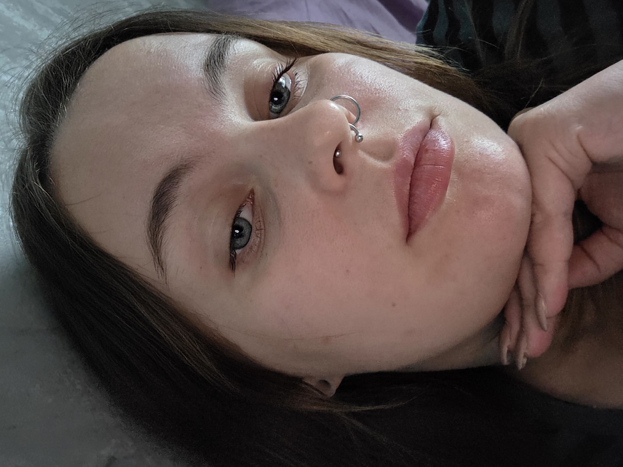 This is a close-up, selfie-style, medium-shot photograph of a young adult Caucasian female. She has fair skin, light brown hair that frames her face, and blue eyes. She is wearing a septum piercing and a nose ring. Her left hand is resting under her chin, with her fingers splayed. The background appears to be indoors, with a dark, textured surface behind her head and a blurred purple surface to her right. There is no indication of the specific location within the city of Minsk, Belarus.