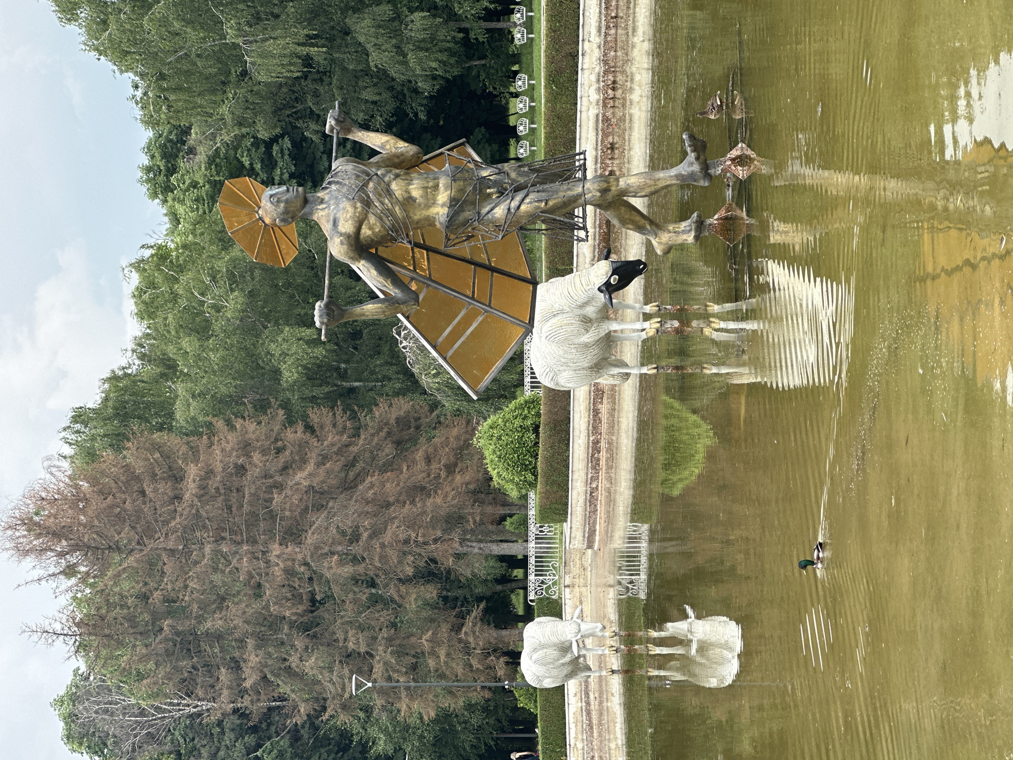 This image depicts a public fountain in Sofia, Bulgaria, featuring a striking sculpture of a golden man carrying a structure over his shoulders, flanked by two white sheep statues. The man's sculpture is abstract, with a sun-like headdress and a woven frame around his body. The sheep statues are realistic, one with a black head and the other with a white head, both standing in the shallow water of the fountain.
In the water of the fountain, a mallard duck swims in the foreground. The fountain's edge is lined with ornate white fencing and manicured bushes. Beyond the fountain, a park setting is visible, with lush green trees and a prominent dead tree with brown foliage. The sky is overcast with scattered clouds. The overall scene captures an artistic display within a natural park environment.
