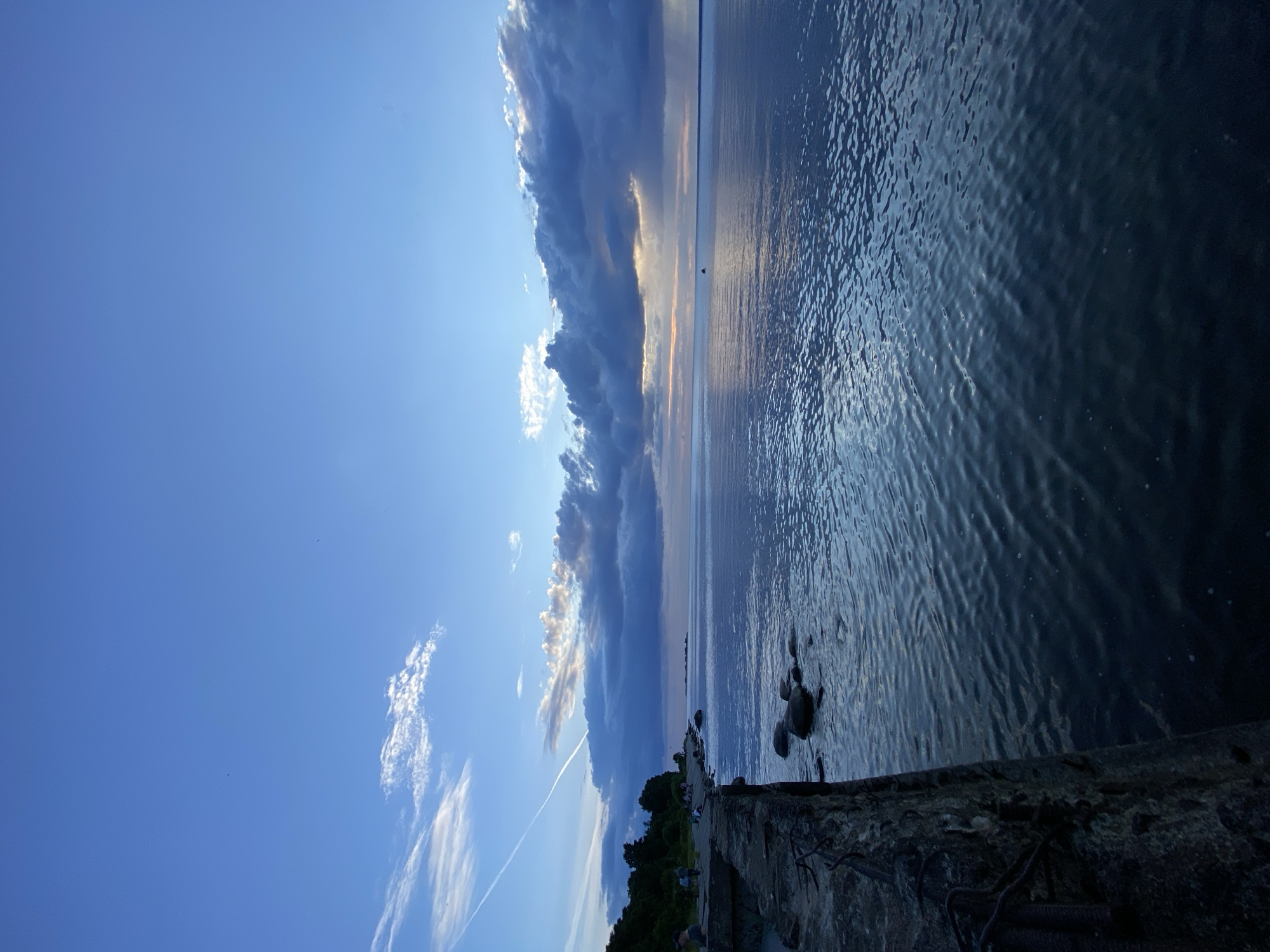The image depicts a wide view of the Gulf of Finland near Sestroretsk, Russia, at dusk. The foreground is dominated by a concrete structure, possibly part of an old pier or breakwater, with visible rebar. To the right of the structure, calm water stretches out, reflecting the blue and grey hues of the sky. The water is slightly rippled, creating patterns of light and shadow.
In the middle ground, along the coastline, a strip of green vegetation lines the shore, interspersed with a few scattered rocks. A small group of people, appearing as silhouettes, are visible walking or sitting along the edge of the water and the concrete structure.
The sky above is a dramatic mix of deep blue and darker, heavy clouds, particularly towards the horizon. A thin, white contrail stretches diagonally across the upper left quadrant of the sky. Sunlight is breaking through the clouds in the distance, casting a warm glow on the horizon. A solitary small boat or buoy is visible on the water in the far distance. The overall atmosphere is serene and tranquil, characteristic of the late evening by the sea.