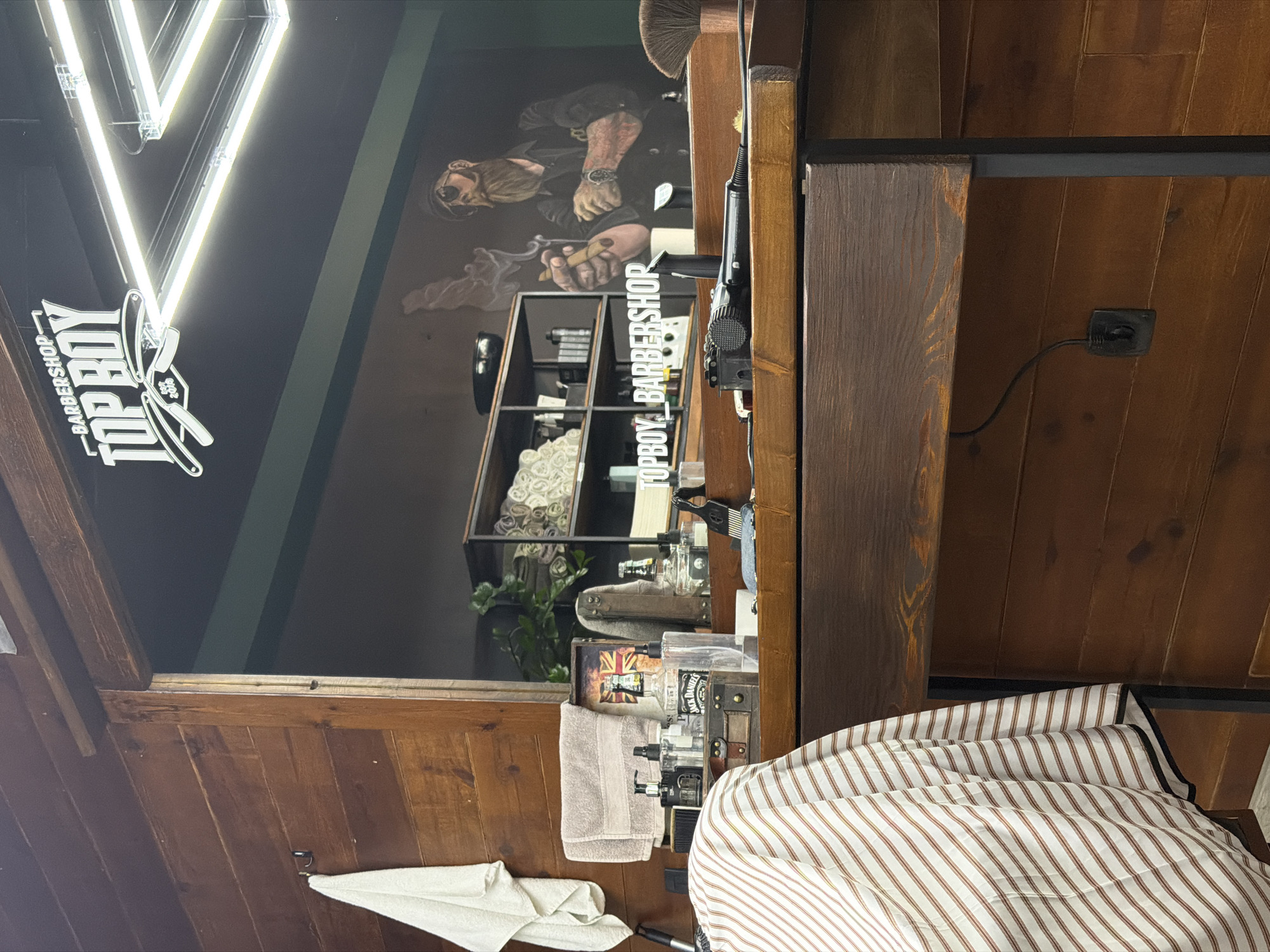 The image shows the interior of the Topboy Barbershop, likely in Budva, Montenegro. The space features a dark gray wall with a mural of a bearded man smoking a cigar. The "Topboy Barbershop" logo is illuminated by an LED sign on the wall. Below the mural is a shelf unit displaying barbering tools. A wooden barber station is adjacent, holding various bottles and a hair trimmer. A white and tan striped barber cape and white towel are draped over the station. The ceiling is wooden, with a black electrical outlet visible. The overall setting is that of a traditionally styled barbershop.