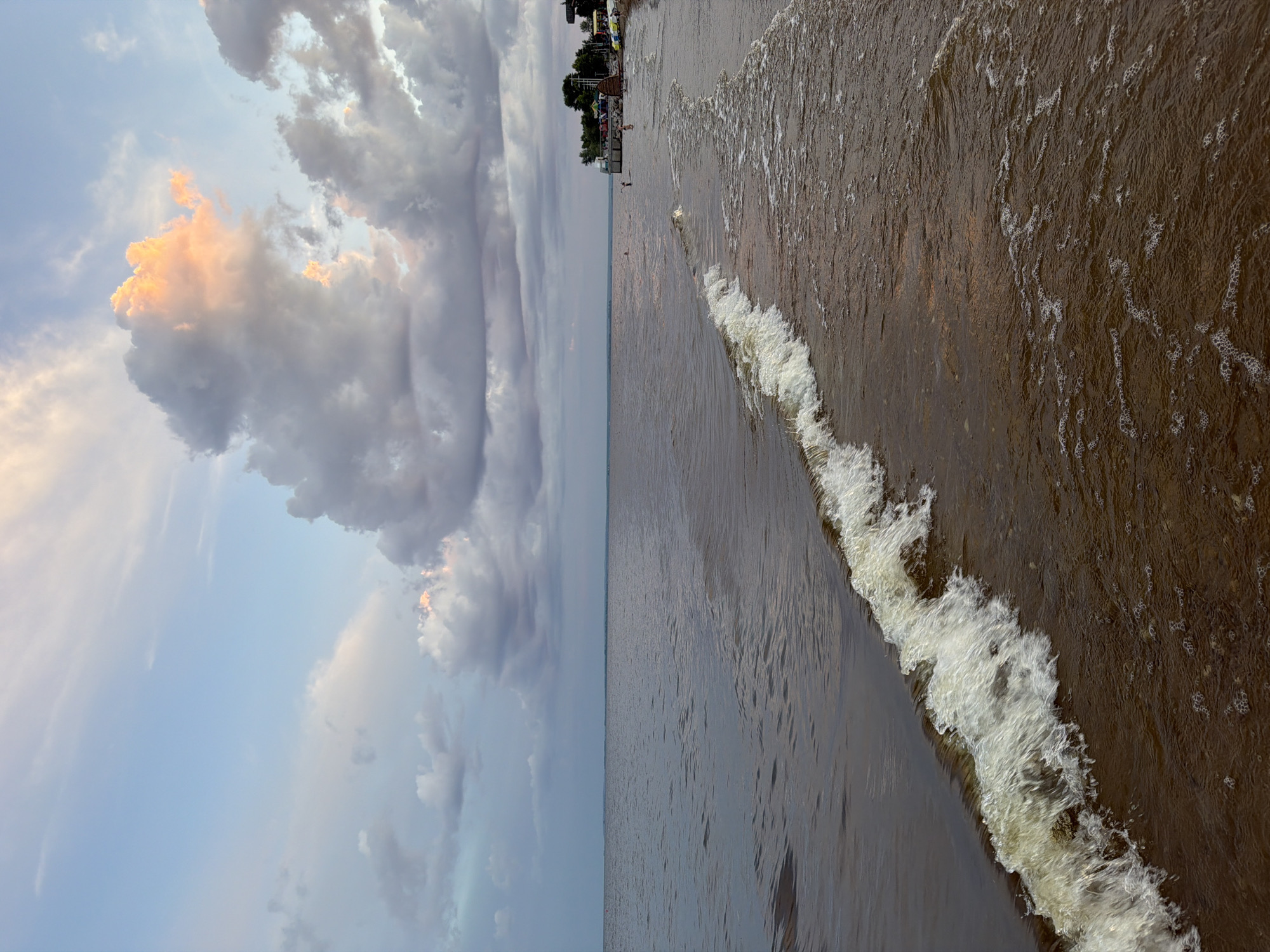 The image depicts a beach scene in Laishevo, Russia. In the foreground, a small wave gently breaks on a sandy shore, with foamy white crests and bubbles trailing in the shallow water. The water is a murky brown color, suggesting sediment or a shallow, silty bottom. In the mid-ground, several people are visible near the water's edge, some appearing to be swimming or wading. A line of trees and buildings, likely part of a resort or town, stretches along the coastline in the distance. The sky above is a mix of pale blue and dramatic clouds, some of which are tinged with pink and orange from the setting sun. The overall impression is one of a tranquil late afternoon or early evening at the beach.