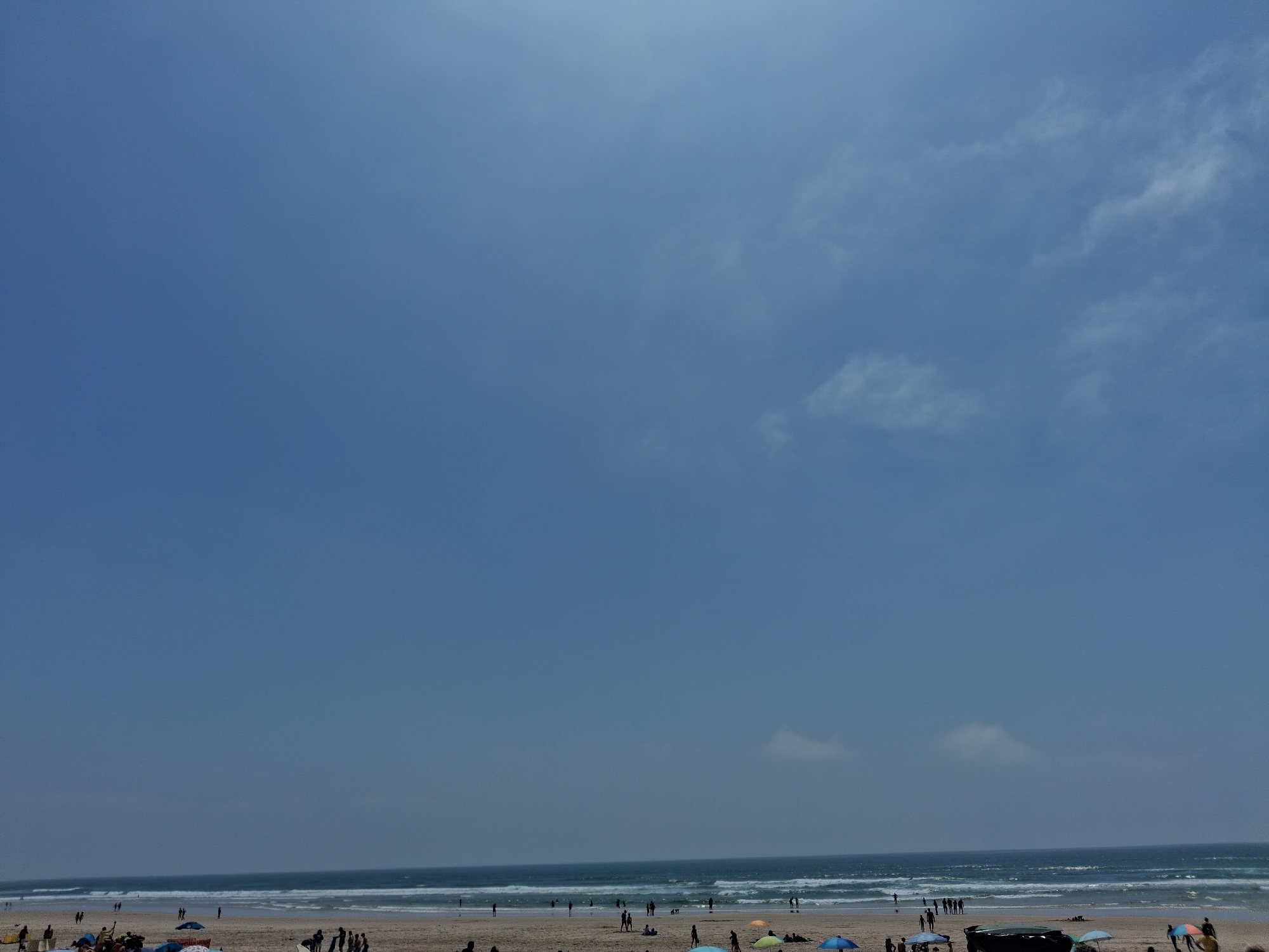 The image shows a beach scene in Figueira da Foz, Portugal. A wide sandy beach is visible at the bottom, occupied by people. The Atlantic Ocean stretches across the middle, with waves breaking near the shore. The sky dominates the upper portion, with shades of blue and scattered clouds. People are walking along the beach and some have set up umbrellas.