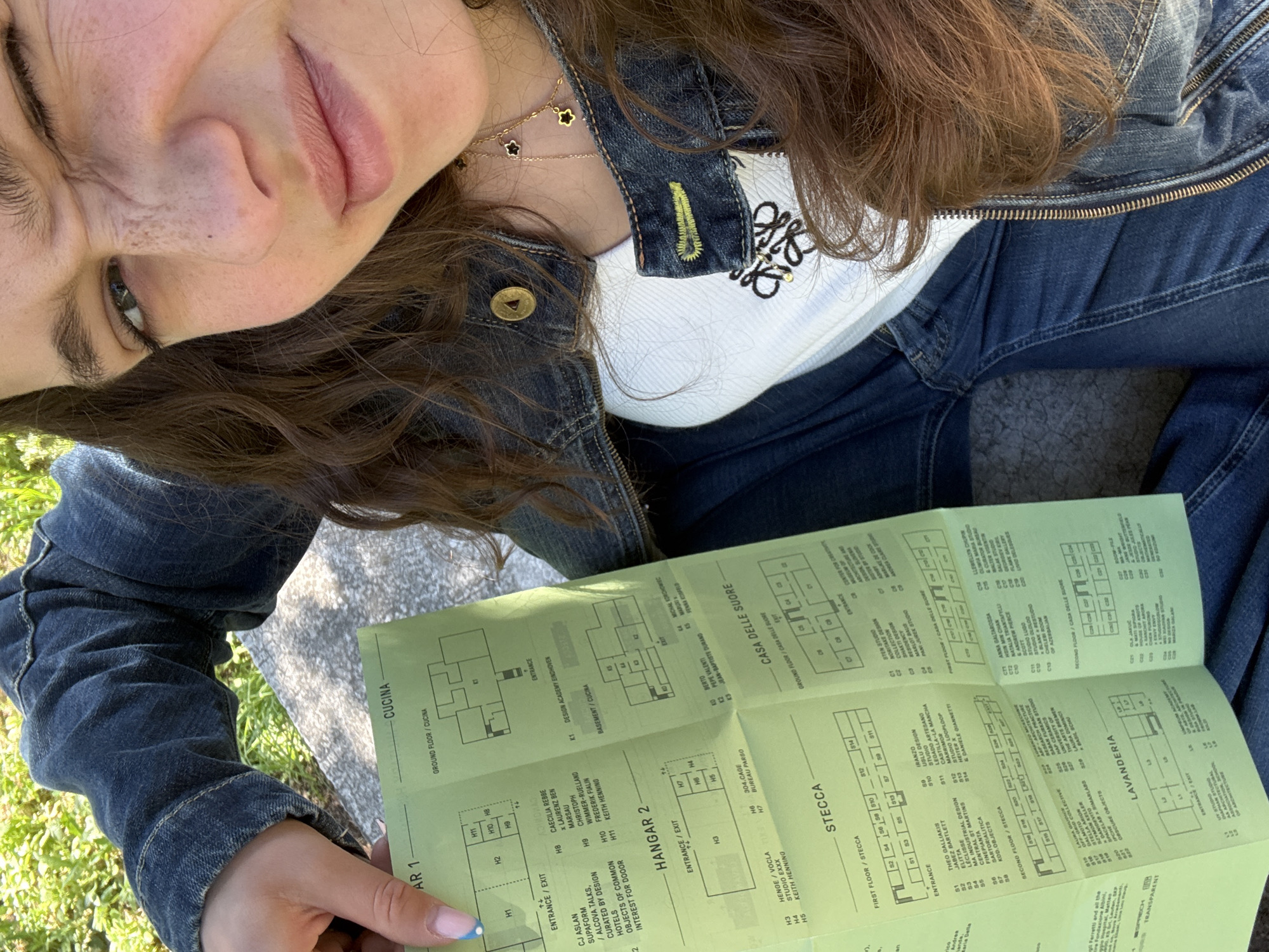 A woman wearing a denim jacket, white shirt, and blue jeans is sitting outside, possibly in a grassy area, looking at the camera. She is holding a green, folded map or brochure in her hands with architectural layouts and text labeled as "CUCINA," "HANGAR 2," "CASA DELLE SUORE," and "STECCA," among other things. The brochure also mentions the "DESIGN ACADEMY EINDHOVEN," and names several designers and studios. The text references "FIRST FLOOR/STECCA" and "SECOND FLOOR/STECCA" possibly corresponding to floor plans on the brochure. The woman is wearing a layered necklace with charms. The surrounding environment appears to be outdoors, with some greenery visible. Given the names of locations and design studios, this image is likely in Milan, Italy.