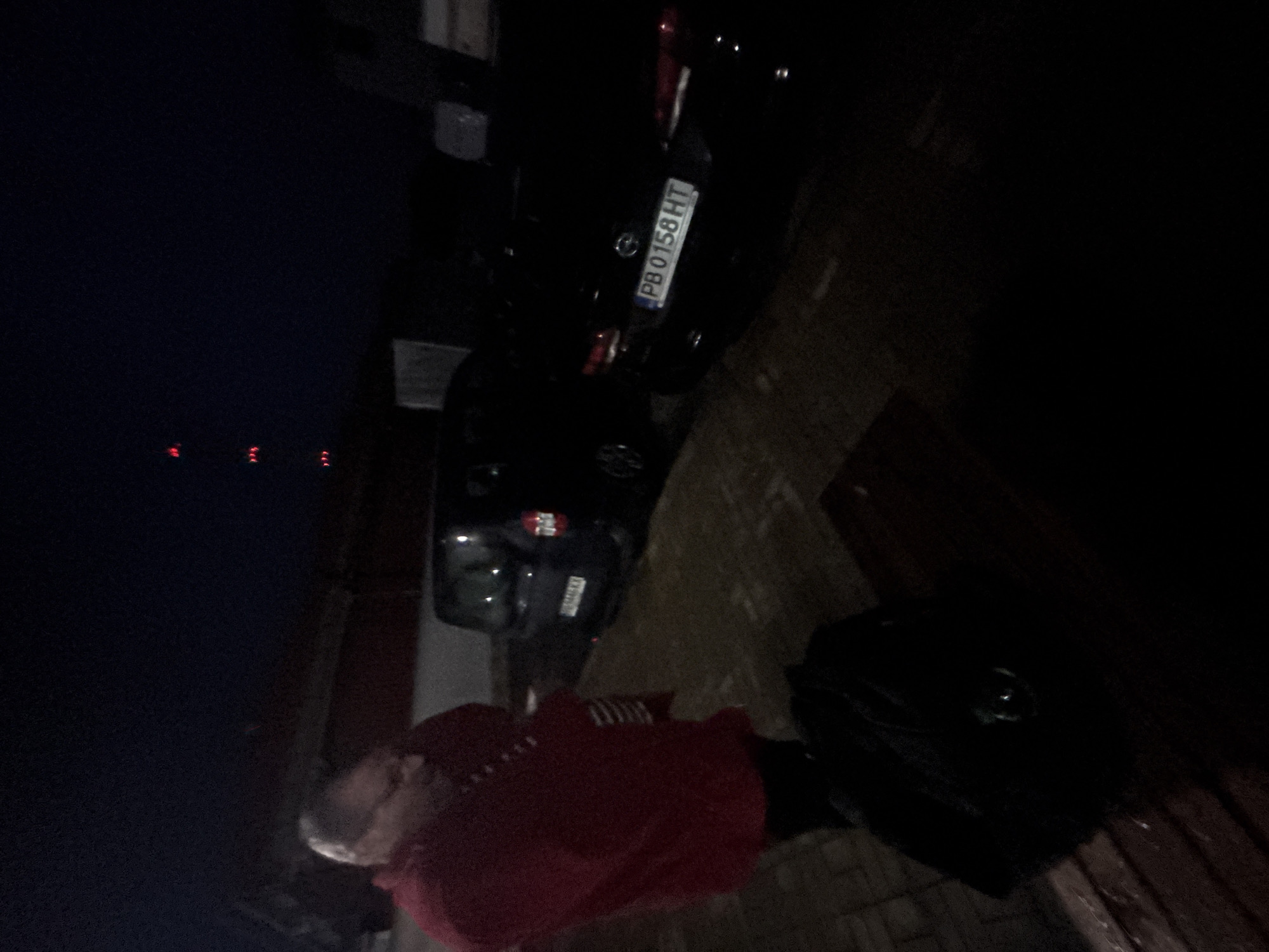 A nighttime scene in Plovdiv, Bulgaria, shows a person in a red shirt standing next to a parked black car. The license plate reads "PB 0158 HT." The environment is dimly lit, with red lights visible in the background. A black bag sits on the ground nearby.