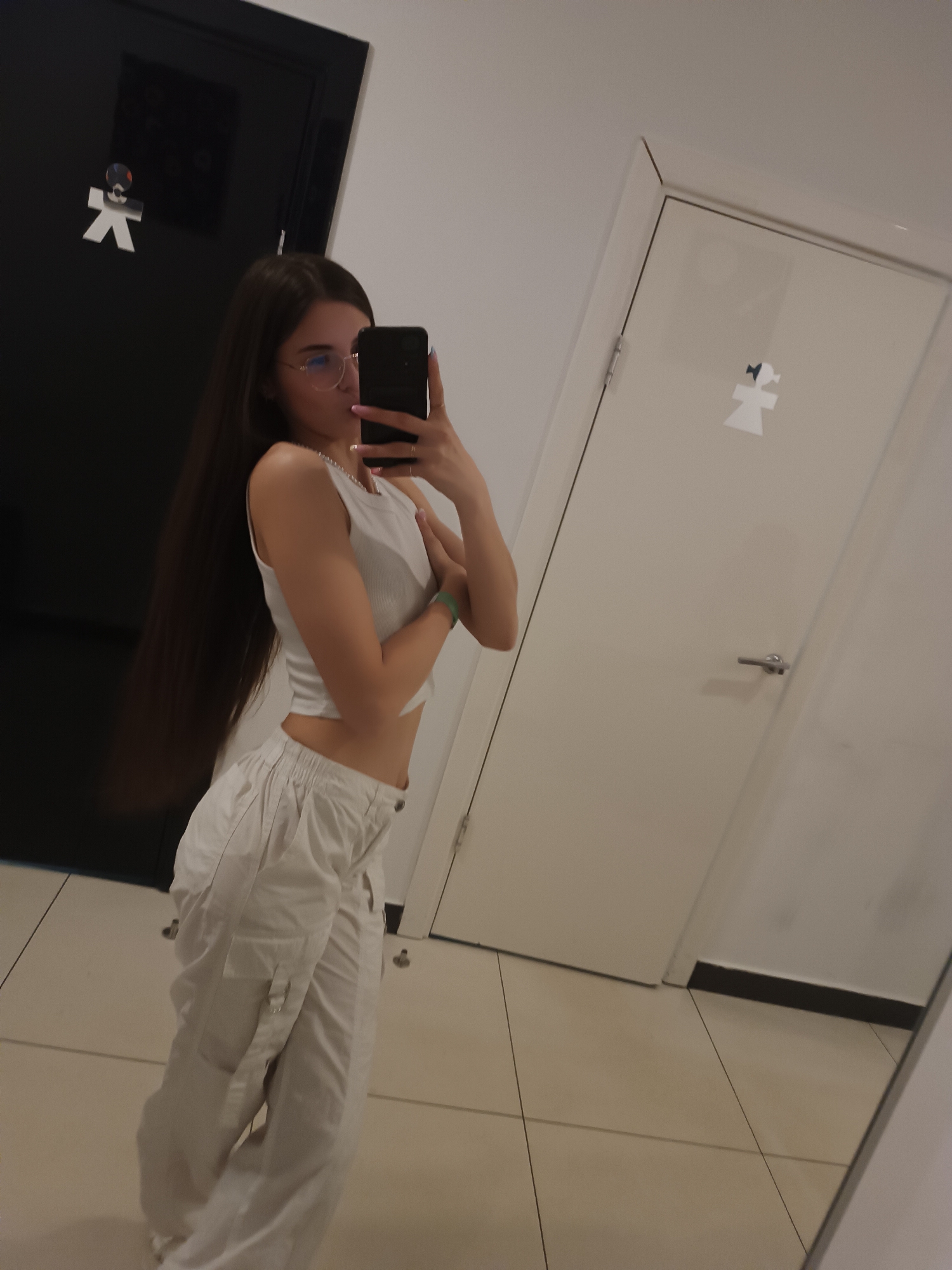A young woman stands in a public restroom, likely in a commercial establishment, taking a mirror selfie. She is dressed in a white tank top and baggy white pants. She is holding her phone in her right hand, obscuring her face from the camera. The restroom has black and white doors with gender symbols. The floor is tiled, and the walls are white. The location is potentially in the city of Chaikovsky, Russia.