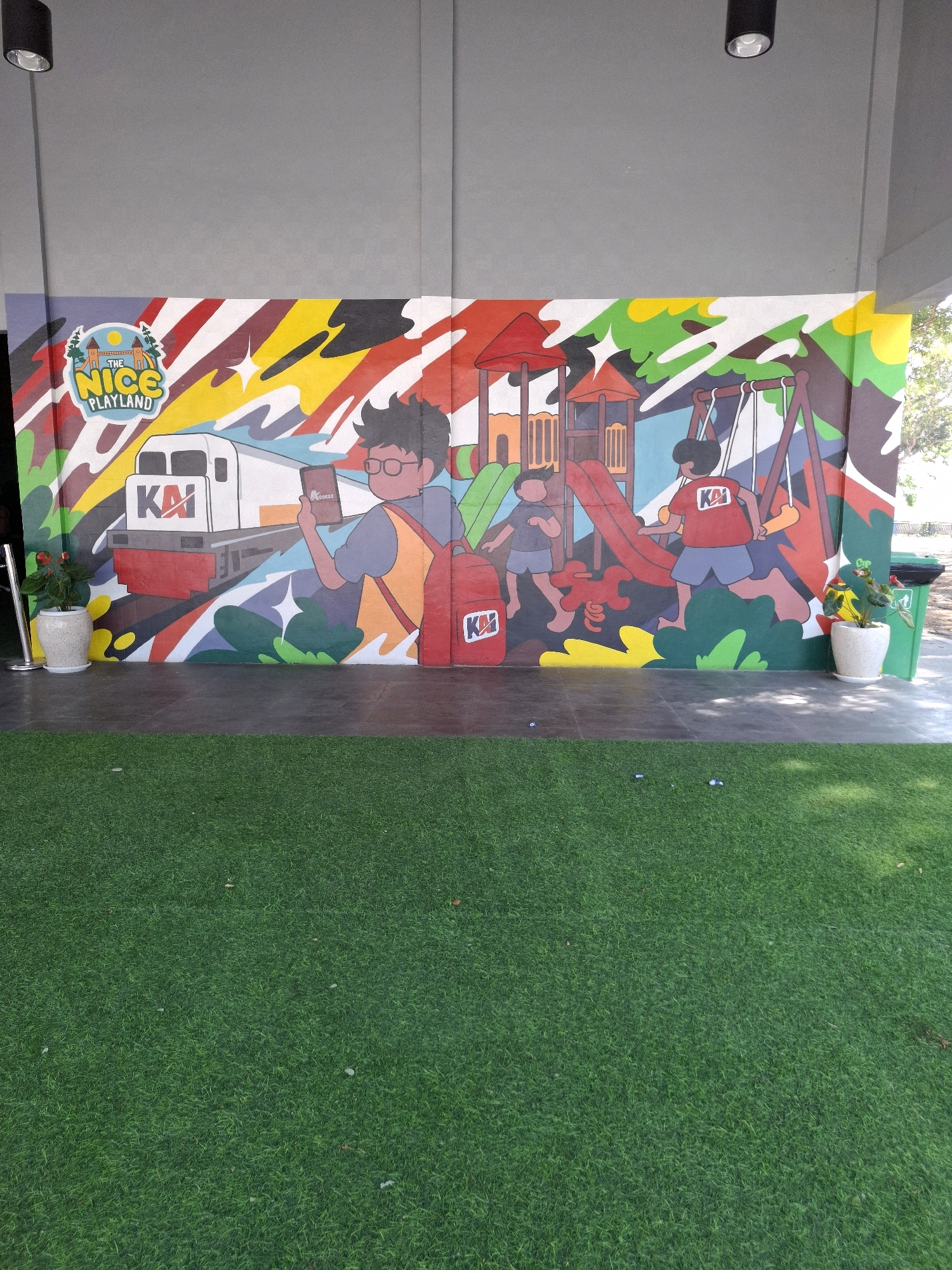 A mural depicting children playing in a playground is painted on a wall in front of a building with a gray facade and a black ceiling. The mural features a train with the logo "KAI", a boy holding a phone, a slide, and swings. Two children are actively engaged in playing. The area in front of the mural is covered with artificial grass. In the foreground, there is a metal barrier with a potted plant and a green trash can. The image is taken in Sidorejo, Indonesia.