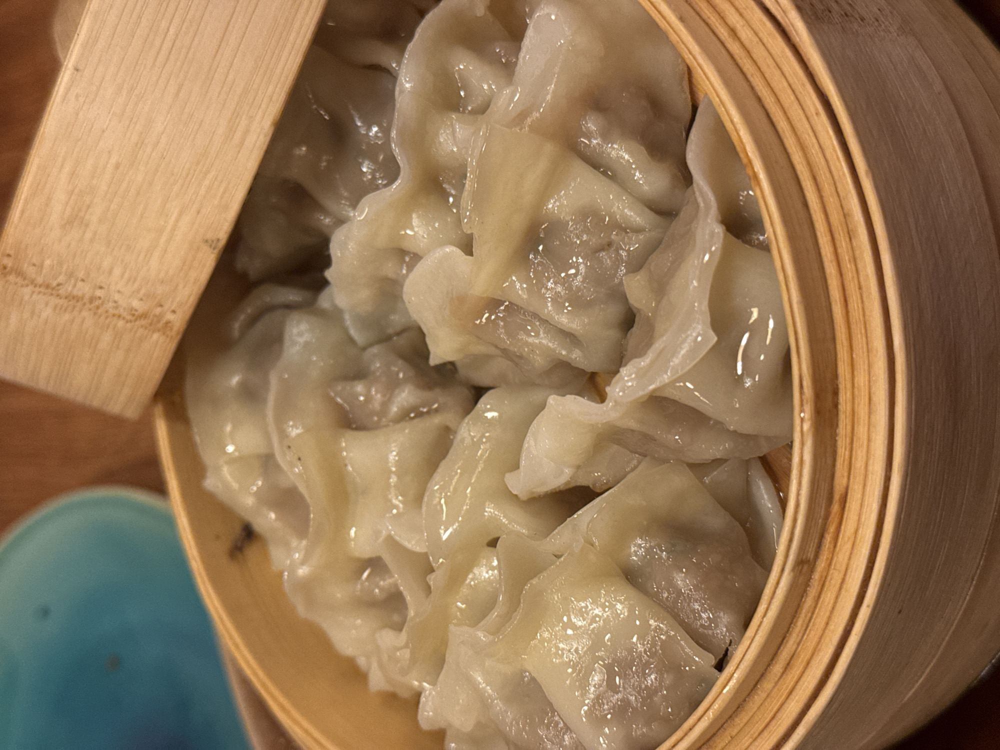 A close-up shot depicts a bamboo steamer filled with dumplings, potentially wontons, with a partially open lid. The translucent wrappers of the dumplings suggest they have been steamed. A blue bowl or saucer is partially visible to the left of the steamer. The scene is likely indoors, with the dumplings potentially at a restaurant in Sofia, Bulgaria. No people are visible.