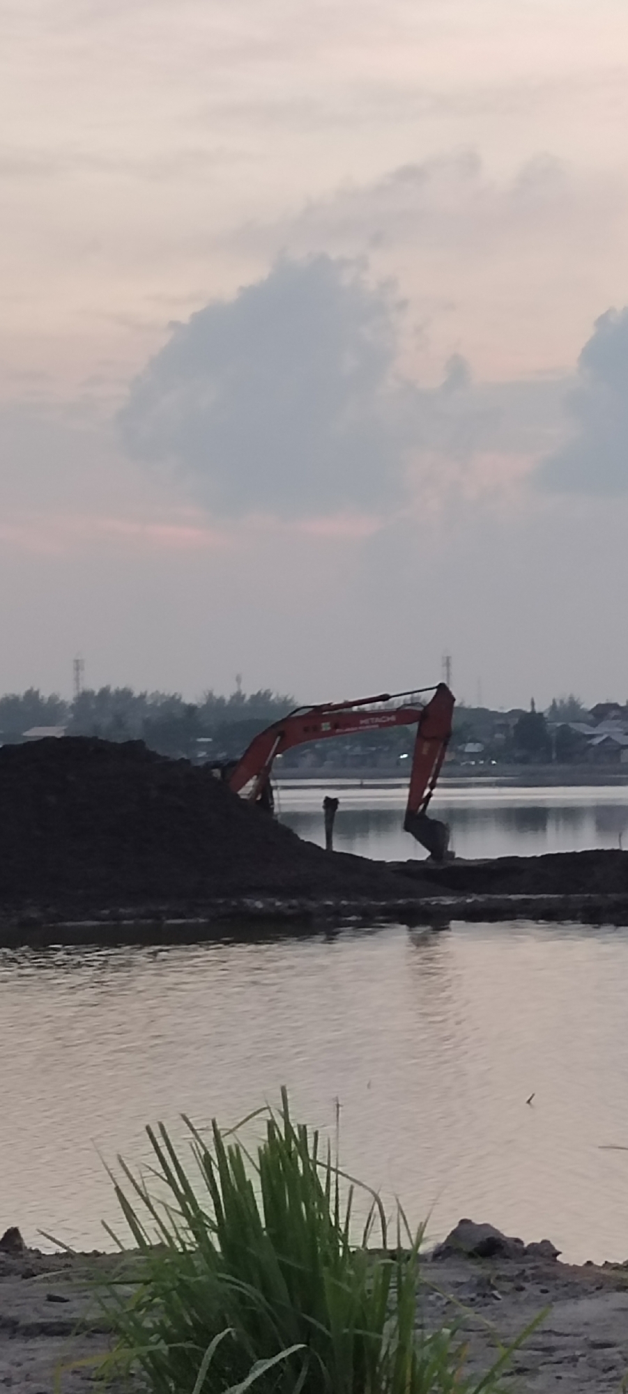 An excavator is visible on the bank of a body of water, with a large pile of dark soil to its left. The excavator is red and features the "HITACHI" logo. The water reflects the overcast sky, which has soft pink and grey hues. In the distance, a treeline with structures can be seen across the water. In the foreground, tall green grass grows on the bank. This scene appears to be a construction or excavation site near a body of water, potentially a river or lake, within the city of Lhokseumawe, Indonesia.