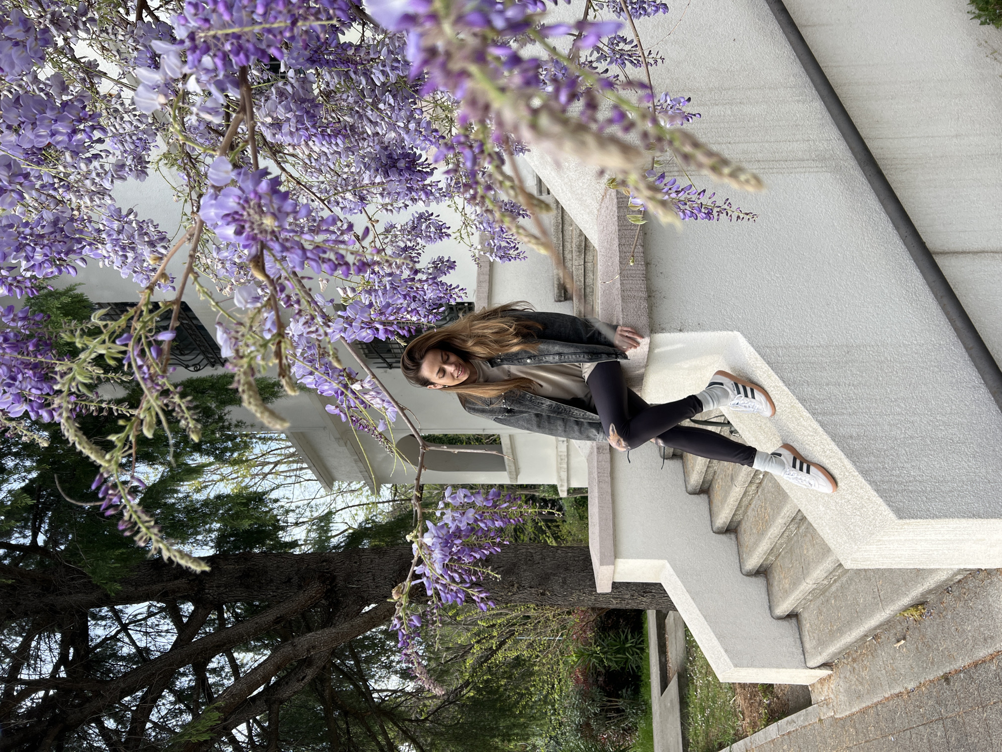 A young woman with long brown hair is sitting on the upper steps of a light-colored concrete staircase. She is wearing black leggings, a beige shirt, a black denim jacket, white socks, and white sneakers with brown soles and black stripes. A large flowering wisteria plant with purple blooms cascades around her and the structure. A mature tree is visible in the background, along with hints of greenery and a white building. The photograph appears to be taken in an outdoor setting.