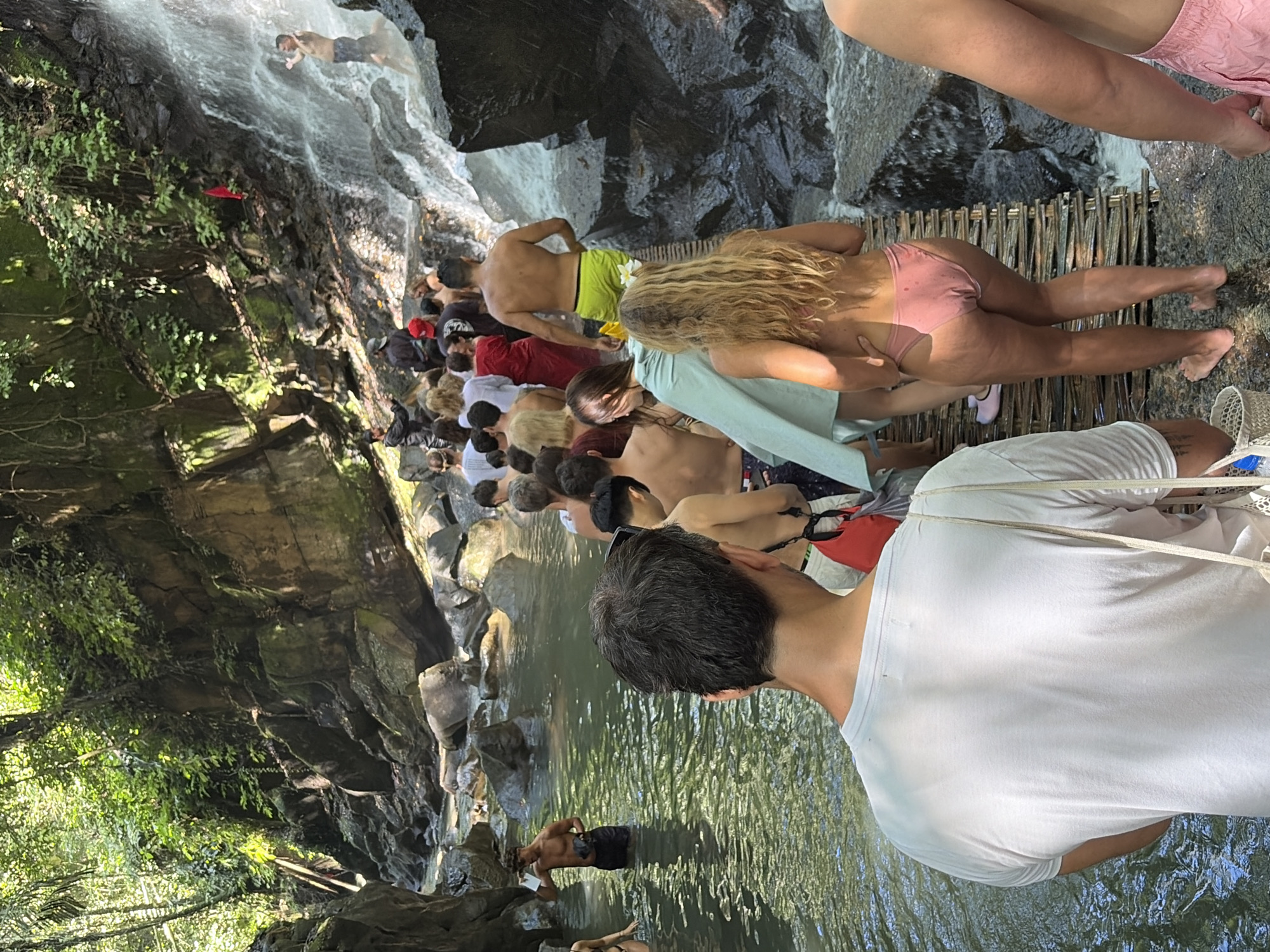The image depicts a group of people in a natural, tropical environment, likely a waterfall or river gorge. In the foreground, a person wearing a white t-shirt is seen from behind, facing away from the camera. To their right, a woman with blonde hair wearing a pink bikini is standing on a wooden walkway. In the middle ground, a procession of people, many shirtless or wearing swimwear, are walking along the wooden walkway and through shallow water. Some are exiting or entering the water. To the far right, a waterfall cascades down rocks, with several individuals visible near and in the water. The environment is lush and green, with trees and vegetation covering the rocky cliffs. The scene appears to be a tourist attraction or a natural swimming spot. This location is likely Tirta Empul Temple or a similar natural spring in Gianyar, Indonesia, known for its spiritual significance and sacred waters.