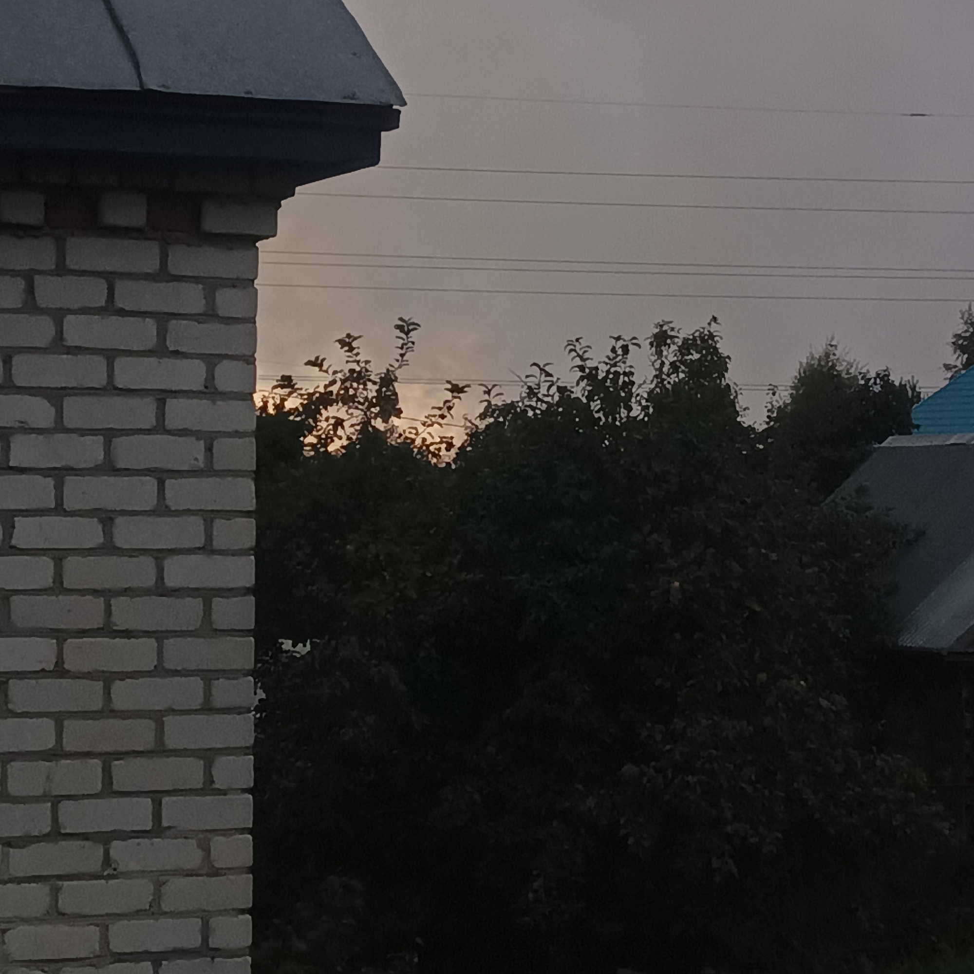 The image depicts an outdoor scene at dusk, likely in Shumerlya, Russia, given the context of the query. In the foreground, the corner of a light-colored brick structure, possibly a building or wall, is visible on the left side, featuring horizontal brick courses. Above this structure, a dark, angled roofline is present. In the background, silhouetted against a pale, overcast sky, is a dense canopy of dark green foliage from trees. Several thin, horizontal power lines crisscross the sky above the trees. To the right, partially obscured by the trees, are the rooftops of other structures, including one with a dark, corrugated roof and another with a bright blue, pitched roof. The overall lighting suggests twilight or a heavily overcast day, with the sun faintly visible through the foliage, creating some bright highlights on the leaves. No people or animals are present in the image.