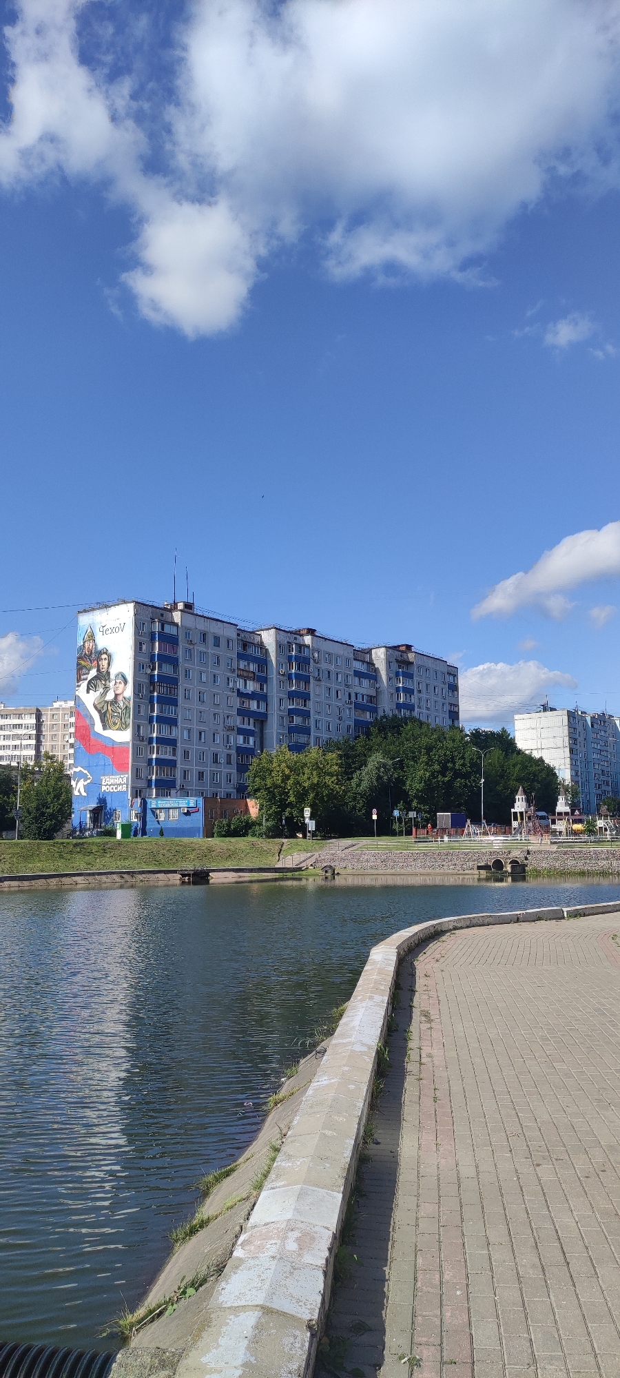 The image depicts a wide, calm body of water in the foreground, bordered by a paved walkway and a sloped concrete embankment with grass. In the background, a multi-story apartment building with a large mural is visible. The mural features portraits of three men in military-style uniforms and the text "ЕДИНАЯ РОССИЯ" (Unified Russia). Adjacent to this building, trees and other residential buildings are present. Further in the distance, on the right side of the frame, a playground structure with small towers is visible, suggesting a public recreational area. The sky is a clear blue with scattered white clouds. The overall scene is a urban landscape with a body of water, residential buildings, and recreational elements.