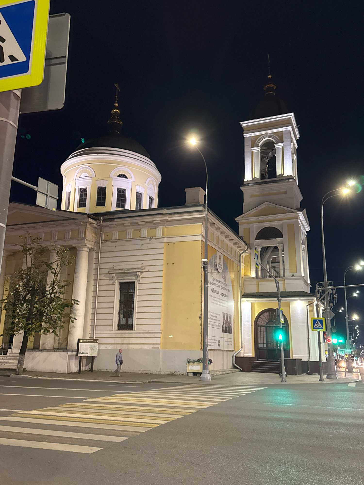 At night, a yellow and white church building with a dome and a bell tower is illuminated in Tver, Russia. A pedestrian crossing is visible in the foreground, with a traffic light showing green. A man in a blue jacket and light-colored pants walks on the sidewalk. A sign with text and images is posted on the church wall. Streetlights and car headlights are visible in the background. A pedestrian crossing sign is mounted on a pole near the church entrance.