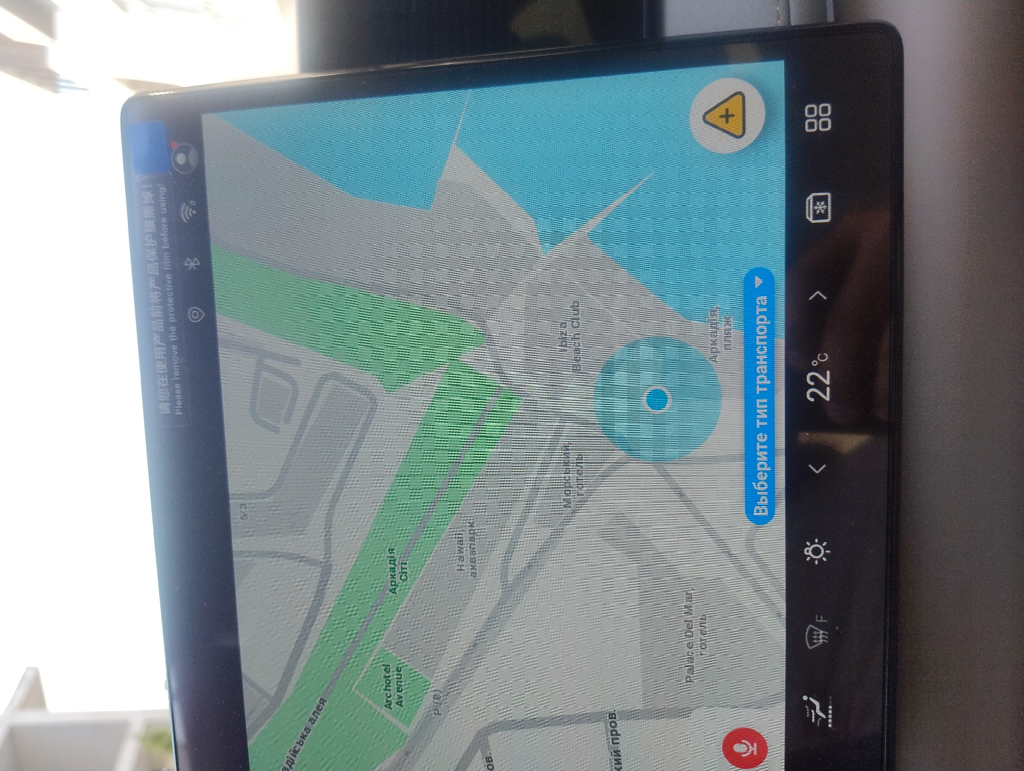 The image shows a close-up of a car's digital navigation screen displaying a map. The screen is reflecting sunlight. Text in both Chinese and English, "请您在使用产品前将产品保护膜撕掉!" and "Please remove the protective film before using!", is visible at the top of the screen, indicating the need to remove a protective film. The map shows a coastal area with green park-like sections and a large blue area representing water. Several location names are visible on the map, including "Archotel Avenue", "Аркадія City", "Hawaii аквапарк", "Морський готель", and "Palace Del Mar hotel". A circular blue target with a white dot is centered on the map, and a yellow warning triangle with a plus sign is in the lower right corner. The bottom of the screen displays a control panel with icons for climate control, a defrost function, brightness adjustment, navigation arrows, temperature reading (22°C), and other system functions. The bottom edge of a car's dashboard is visible.