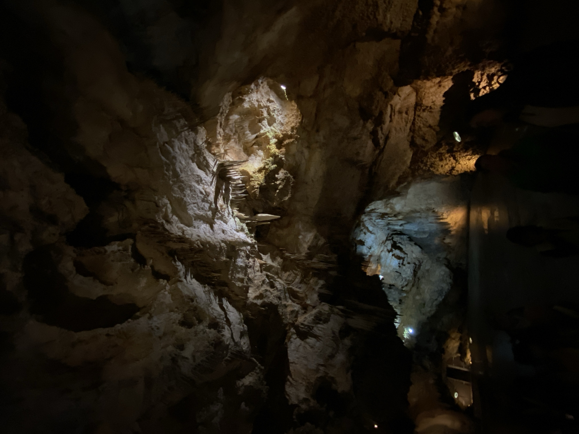 The image shows a dimly lit cave interior in Lipa Dobrska, Montenegro. The cave walls are rugged and uneven, with various rock formations. A series of steps have been constructed on the rock face, leading upwards. Some parts of the cave are illuminated by artificial lights, revealing the texture of the stone. To the right, a reflective surface, likely a glass barrier, is partially visible, obscuring details beyond.