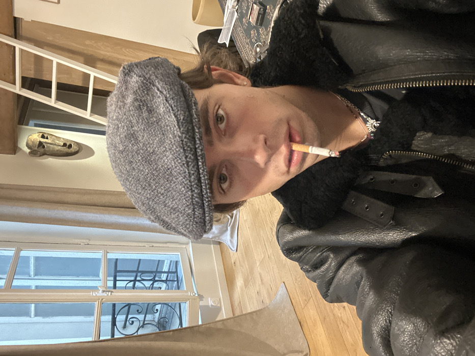 A young male individual is depicted indoors, looking directly at the viewer. He wears a grey tweed flat cap, a black leather jacket with a black shearling collar and lining, and multiple silver-colored necklaces. A lit cigarette, with an active ember, is held between his lips. The background reveals a room with light wooden floorboards and light-colored walls. To the left, a white-framed French window with multiple panes is partially open, exposing a wrought-iron balcony railing and the facade of an adjacent building, indicative of Parisian architecture. Light-colored curtains are drawn to the sides of the window. In the upper right background, a white wooden ladder leans against a wall beneath a visible wooden beam. A stylized, golden-hued mask is mounted on the wall below the beam. On a surface in the lower right, a patterned textile and various objects, including what appears to be a cigarette pack, are visible. The setting is a residential interior in Paris, France.