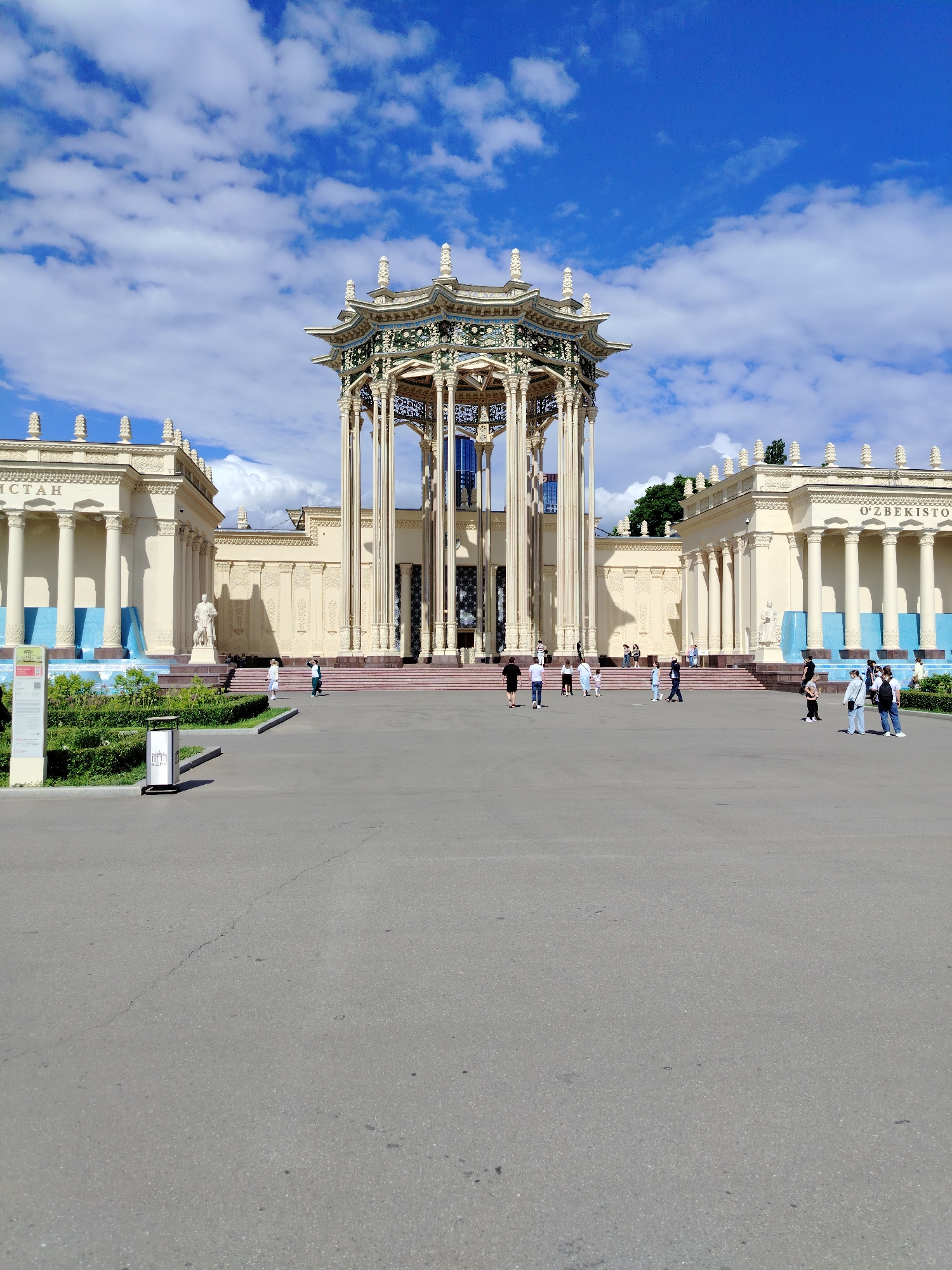 This is a medium shot photograph taken outdoors on a sunny day with scattered clouds, likely in the afternoon. The scene depicts a large, ornate pavilion with a central, multi-tiered roof structure supported by tall, decorative columns. This pavilion is flanked by two symmetrical, cream-colored buildings with classical architectural elements, including rows of columns and decorative friezes. The building on the left has the word "ИСТАН" visible, and the building on the right has "O'ZBEKISTO" visible, suggesting these are pavilions representing republics of the former Soviet Union, likely at an exhibition.
In front of the pavilion is a wide, paved courtyard where several people are walking. They are casually dressed, and their presence suggests a public space that is open and accessible. Some individuals are walking individually, while others are in small groups.
The foreground shows the paved surface of the courtyard, with a slight crack visible in the asphalt. To the left of the frame, there is a manicured green hedge and a tall information stand. Behind the hedges, blue tiled fountains are visible along the base of the flanking buildings.
The overall impression is one of a historical or cultural exhibition site, perhaps part of a larger park or complex, with visitors enjoying the architecture and pleasant weather. The bright sunlight and clear blue sky contribute to a feeling of openness and a positive atmosphere.