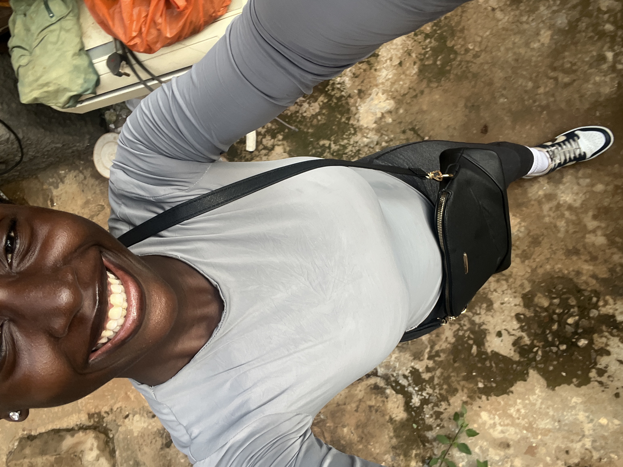 A dark-skinned woman with a wide smile and a stud earring is taking a selfie in Abuja, Nigeria. She is wearing a light gray long-sleeved shirt, dark leggings, white socks, and black and white sneakers. A black crossbody bag with gold hardware hangs across her chest. The background appears to be an outdoor, slightly unkempt area with dirt ground and some discarded items, including a green bag and an orange plastic bag, on a white surface to the right.