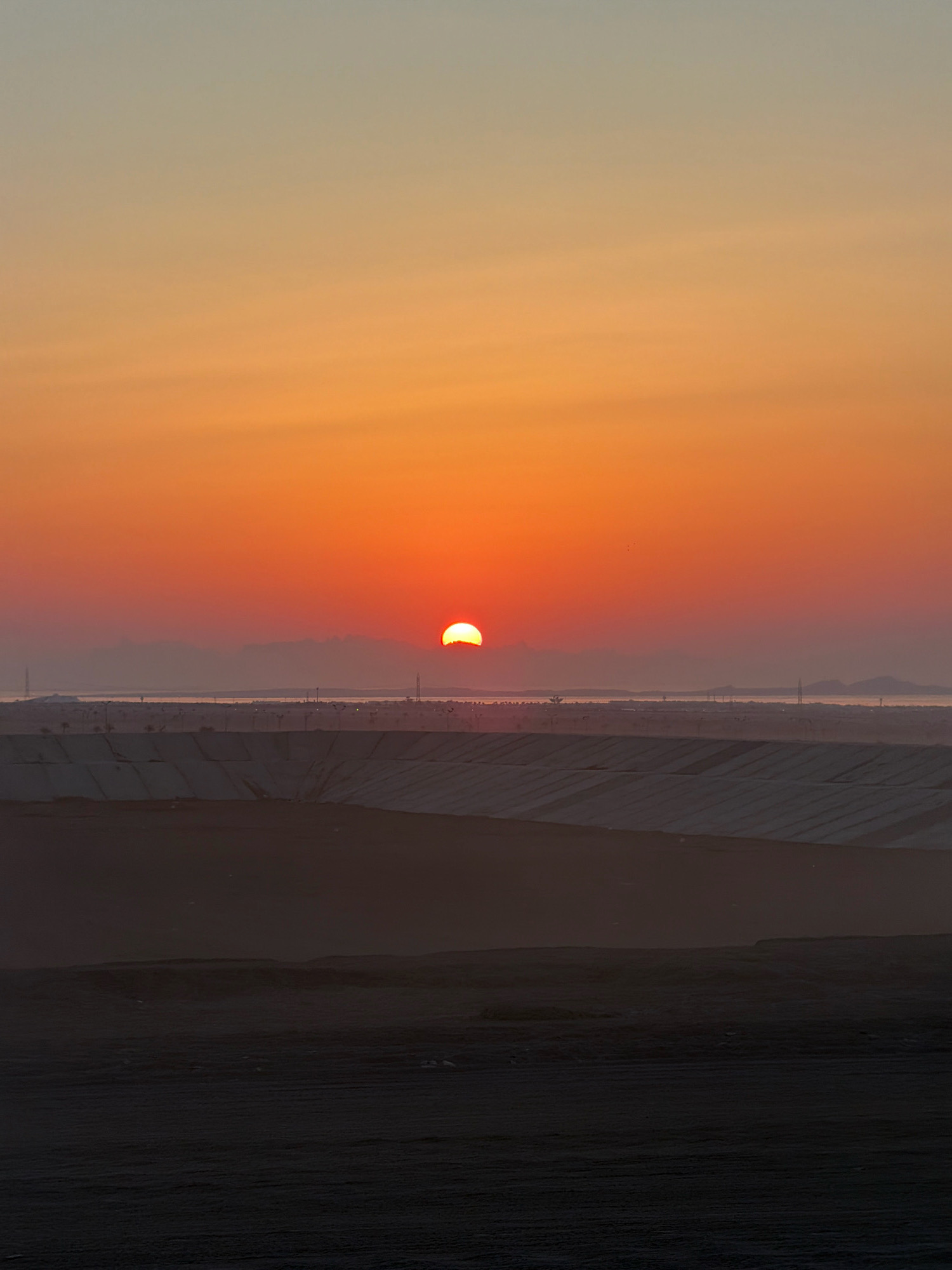 A sunset is depicted over a landscape in Sharm El Sheikh, Egypt. The sun is a bright orange orb, partially obscured by the horizon, casting a warm glow across the sky. The sky transitions from a pale blue at the top to vibrant shades of orange and red near the horizon. In the mid-ground, a body of water is visible, with distant landmasses and structures on the far shore. In the foreground, there are layers of arid land, with some man-made structures visible, potentially part of infrastructure or development projects. The overall impression is one of a vast, open environment under a colorful sky.