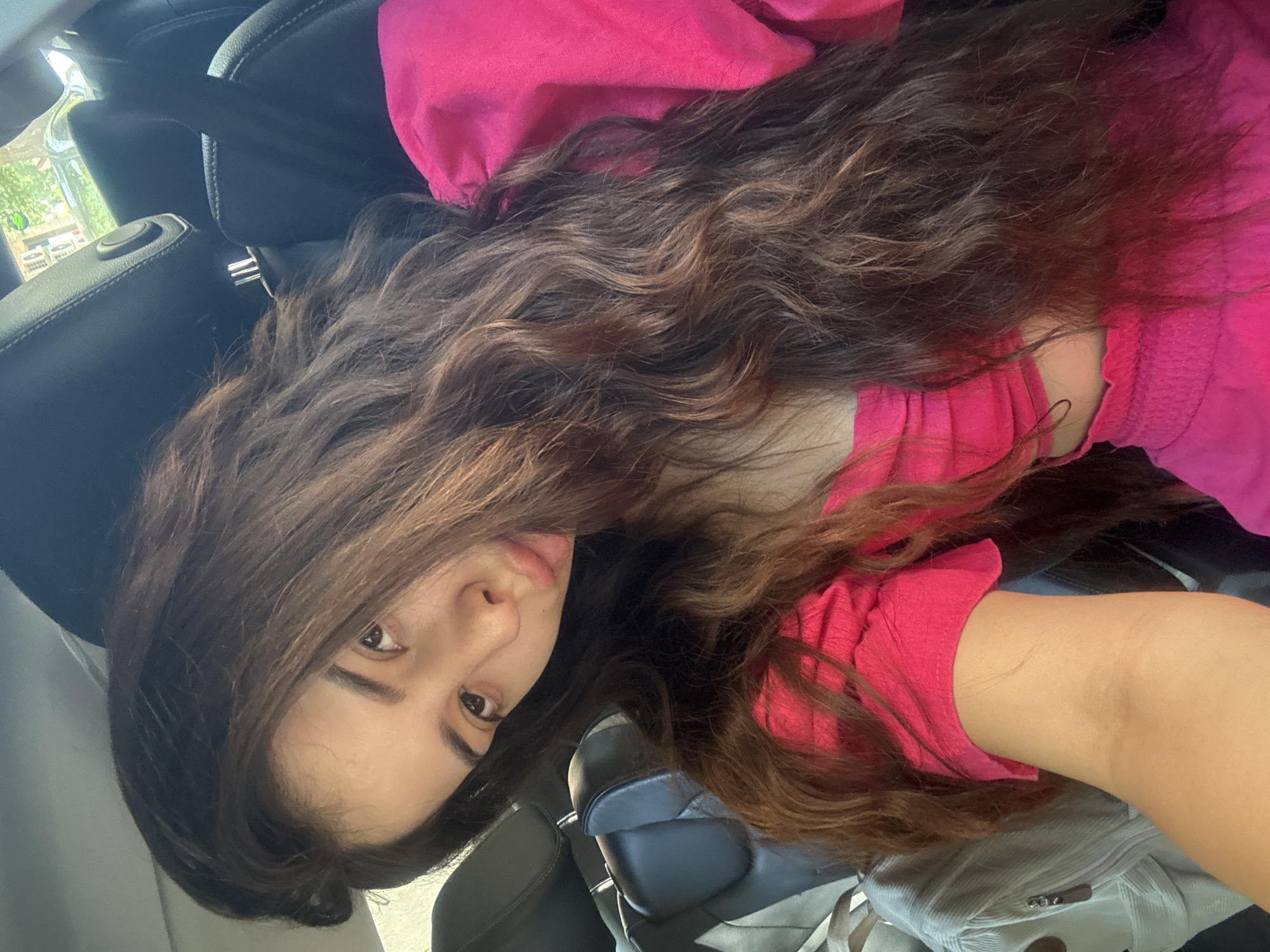 A young woman with long, dark, wavy hair is seen in a car, likely as a passenger. She is wearing a bright pink, off-the-shoulder top and matching pink bottoms. Her head is tilted, and she is looking at the camera with a slight pout. The background shows the interior of a car, including black leather seats and headrests, with a glimpse of greenery and buildings visible through the car window, suggesting an urban environment. The exact location in Plovdiv, Bulgaria, cannot be determined from the image alone.