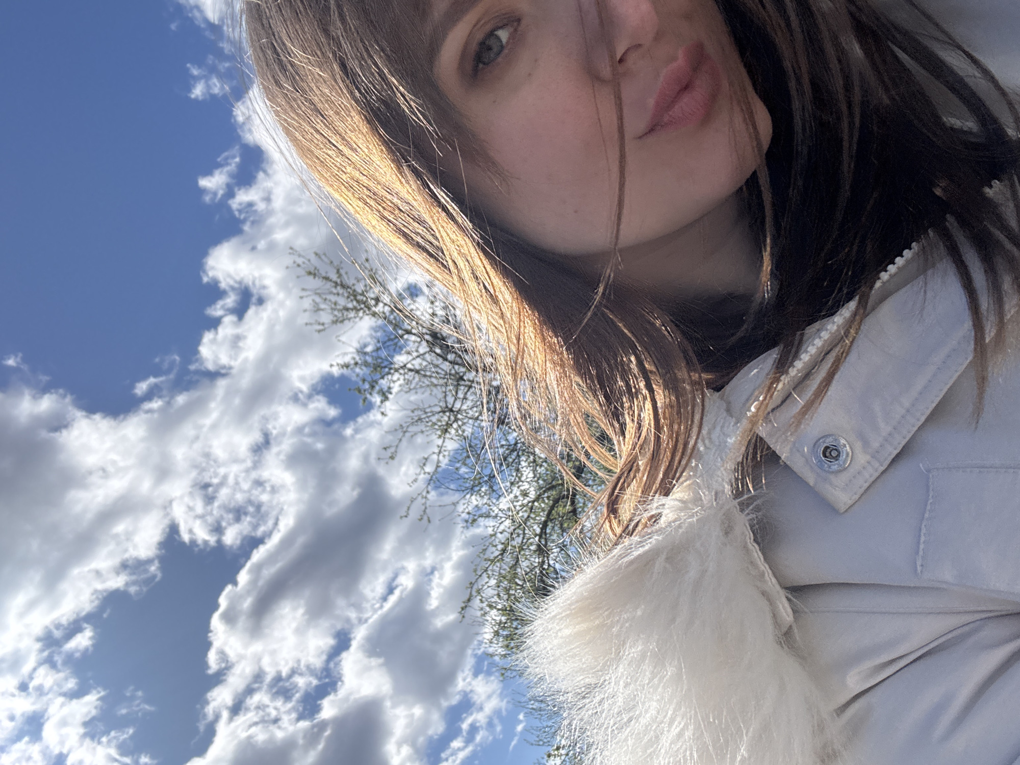 A young woman is pictured in an outdoor setting. She has brown hair, fair skin, and appears to be wearing a light-colored jacket with a fur collar. The background includes a blue sky with scattered white clouds. Some tree branches are visible. The image is angled upward towards the sky. Location is the city of Chaikovsky, Russia.
