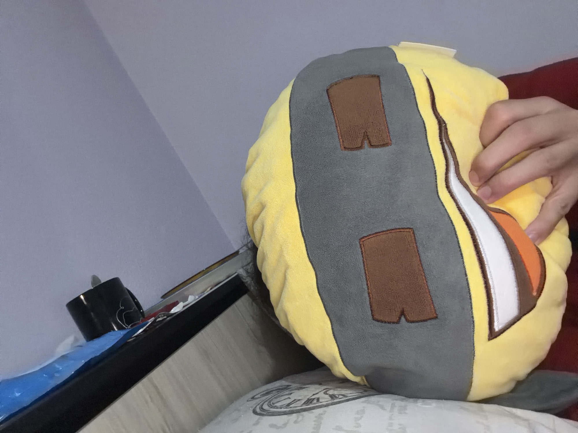 The image presents a close-up view of a yellow and grey plush toy, seemingly a pillow or cushion, with a cartoonish design. The toy is predominantly yellow with grey elements, featuring brown rectangular patches reminiscent of windows or compartments. A hand is visible, holding or manipulating the toy. The toy rests on a patterned pillow or cushion, suggesting a domestic setting. In the background, a small portion of a wall is visible, as well as the edge of a table with objects, including a black mug.
The location is not discernible from the visual elements within the image.