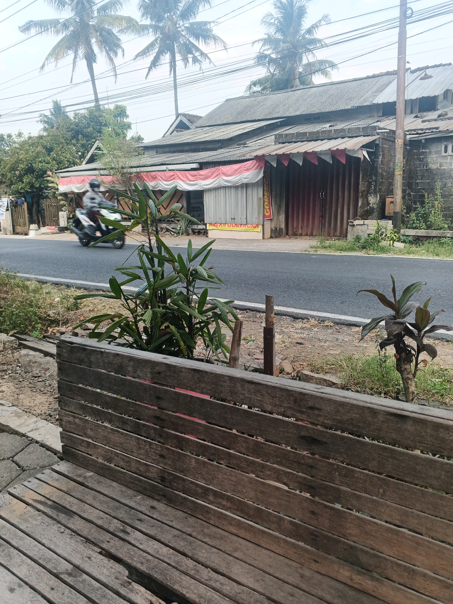 The image depicts a street scene in Ungaran, Indonesia. In the foreground, a weathered wooden bench or fence is visible, constructed from rough-hewn planks. Beyond this, a paved road runs horizontally across the frame. On the opposite side of the road, a building with a corrugated metal roof and wooden shutters is situated. A red and white striped awning stretches across the front of the building, which appears to be a shop or food stall. A motorcycle with a rider wearing a helmet is moving along the road, creating a blur of motion. Palm trees are visible in the background, suggesting a tropical or subtropical climate. The overall scene is overcast, contributing to a muted color palette.