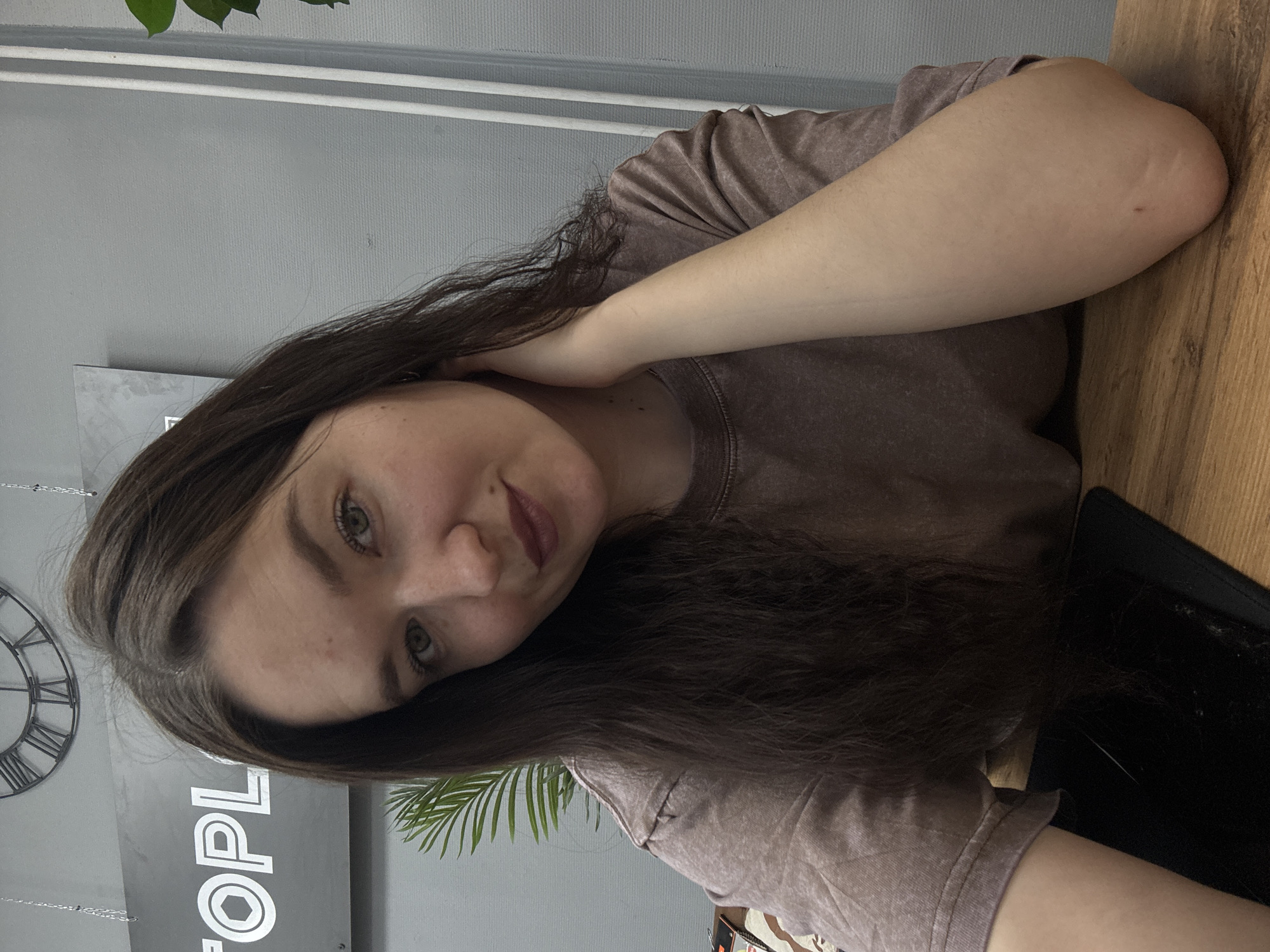 A woman with long, dark hair and fair skin is at a table in a cafe. The woman is wearing a brown t-shirt. A clock and a sign are in the background. The sign has "People" written on it. There are also some plants around. The table is wooden. The woman is resting her arm on the table. The picture was taken indoors during daytime. There is no indication of the image being in the city of Chaikovsky, Russia.