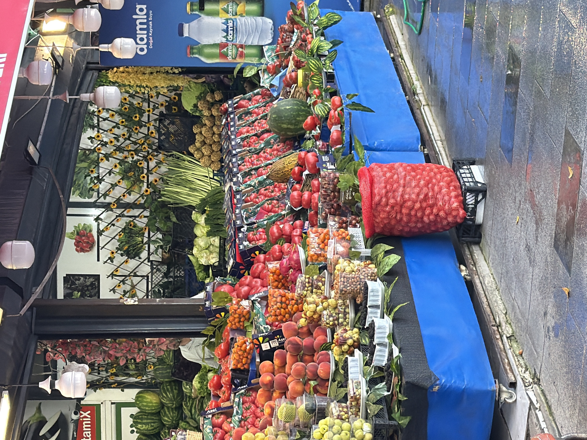 A fruit and vegetable stall is displayed in an outdoor market in Kadıköy, Turkey. The stall is laden with various produce, including watermelons, peaches, tomatoes, plums, grapes, and what appear to be nuts or small round fruits in containers. Several large, red mesh bags filled with produce are also visible. Behind the produce, decorative backdrops of sunflowers and foliage are displayed. Several bottles of "Damla" mineral water are placed on a blue surface to the right of the stall. The ground in front of the stall is wet, suggesting recent rain, and is paved with large, grey tiles. A person is partially visible behind the display, wearing a light-colored shirt. The overall scene depicts a vibrant and bustling market environment.