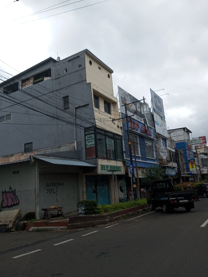 This image depicts a street scene in the city of Vaatharawattai, Sri Lanka, under an overcast sky. The view is of a street, looking along a two-lane asphalt road with a double-dashed white centerline. On the right side of the street, a row of multi-story commercial buildings is visible.
The leftmost building is a three-story structure predominantly grey. Its ground floor features a closed shop with a corrugated metal awning; a rolled-down security door displays white text reading 'SUPREMASI SIPIL'. Pink and purple graffiti, resembling 'JUH', is present on the lower left wall. In front of this shop, a wooden bench or table supports a small, indistinct brown object.
Adjacent to the grey building, another multi-story structure has a cream-colored upper section and a light grey lower section. The ground floor retail space features large glass windows displaying multiple signs with the text 'DIJUAL' (For Sale), and a green fascia sign reads 'ELY DIESEL'. To its immediate right, a closed shop displays light blue graffiti, 'PALADA', on its rolled-down security door.
Further down the street, a blue multi-story building is prominent, adorned with several large advertising billboards on its upper facade, one featuring a stylized 'GO' logo. A tall street pole with an ornate, caged lantern fixture and numerous overhead electrical wires is positioned in front of this blue building. Additional commercial structures with various signs extend into the distance.
A dark-colored pickup truck is parked on the right-hand shoulder of the road, facing away from the viewer. No discernible individuals are present within the visible area.