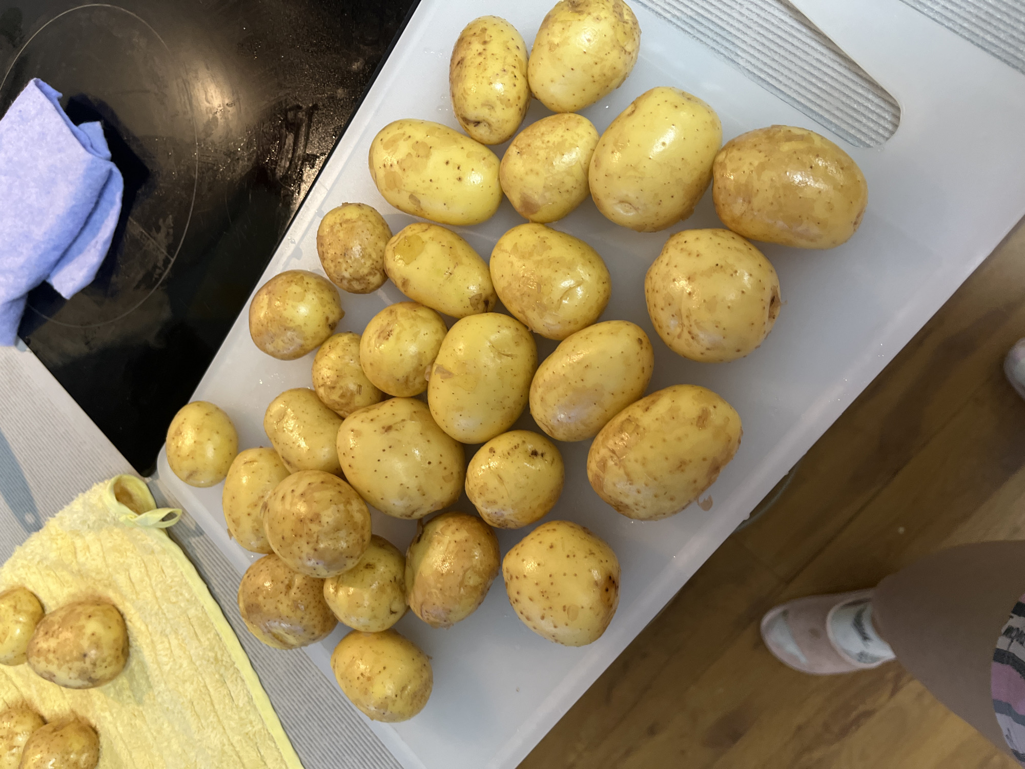 The image depicts a domestic kitchen scene, likely in Sochi, Russia, focusing on the preparation of food. The main subjects are numerous small, yellow-skinned potatoes, which appear freshly washed. Most of these potatoes are spread across a white cutting board, positioned centrally and to the right. A smaller cluster of potatoes rests on a yellow kitchen towel in the bottom left corner of the frame.
The setting includes a black stovetop or induction hob in the upper left, upon which a folded blue cloth is placed. The white cutting board is situated on a light-colored kitchen counter. In the bottom right corner, a person's lower leg and foot are visible, wearing a pink slipper and a striped sock, indicating a casual home environment.
The activity taking place is the initial stage of preparing potatoes for cooking, specifically after they have been washed and are either drying or awaiting further processing. The potatoes show natural variations in size and some small dark specks on their skin. The lighting suggests an indoor setting, but no specific time of day or weather conditions can be determined. There is no legible text visible within the image. The overall impression is one of practical kitchen work.