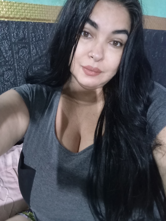 A woman with long, dark hair is centered in the frame. She has light skin, brown eyes, and wears lipstick. She is dressed in a gray t-shirt with a v-neck. The background includes a patterned headboard and bed linens. The lighting appears to be indoors. The image is a close-up shot.