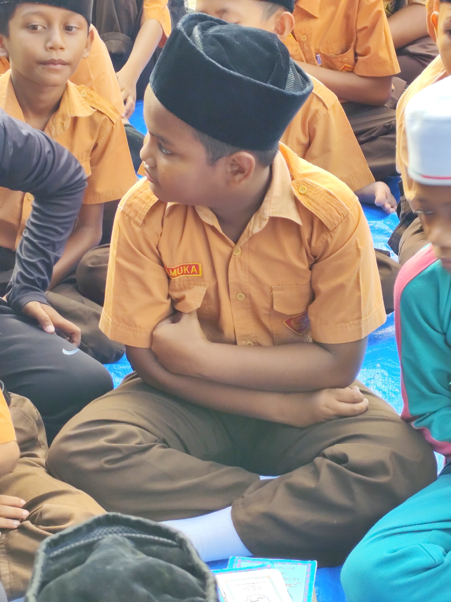The image shows a group of young boys, presumably students, sitting on a blue tarp. They are all wearing similar orange uniforms and dark caps. The boy in the foreground is looking to his left with his arms crossed. Another boy to his right is also visible, wearing a turquoise shirt with pink accents. The overall setting appears to be outdoors, possibly in a schoolyard or a community gathering space. There are books or papers scattered on the tarp in front of the boy in the foreground. The lighting suggests it is daytime.