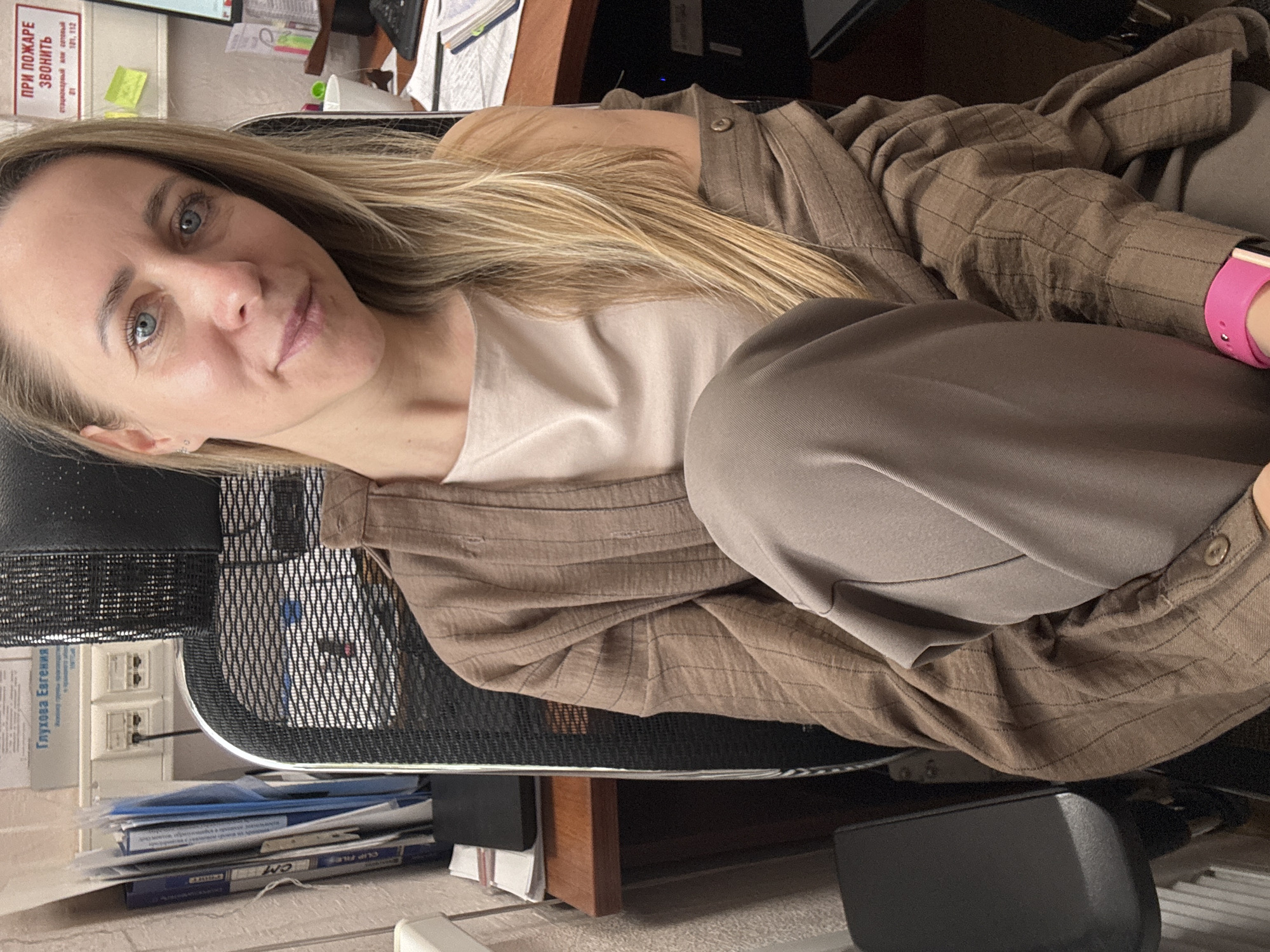 The image shows a woman sitting in an office chair. She has blonde hair and blue eyes, and she is wearing a beige shirt and a brown striped jacket. She has a pink smart watch on her wrist. The background shows an office environment with papers and electrical outlets. The location is in Chaikovsky, Russia, based on the Russian text visible in the image.