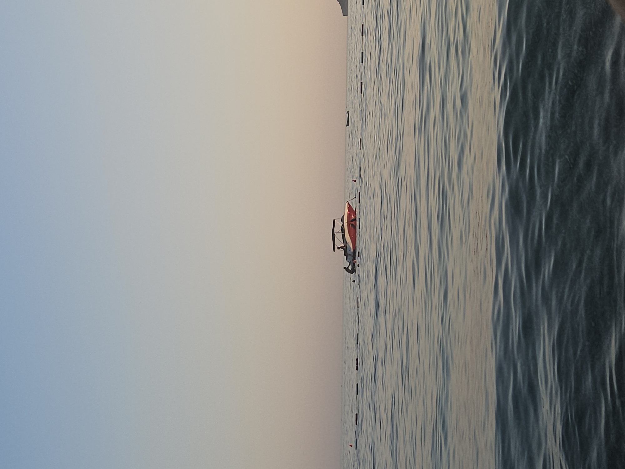 A speedboat with an orange and white hull is moored in the sea, with two people visible on board. In the background, a buoy line stretches across the water. The sky is a pale gradient of blue and peach, indicating either dawn or dusk. The water is calm, with gentle ripples reflecting the sky's colors. The scene suggests a peaceful moment at sea, possibly near a beach or coastline.