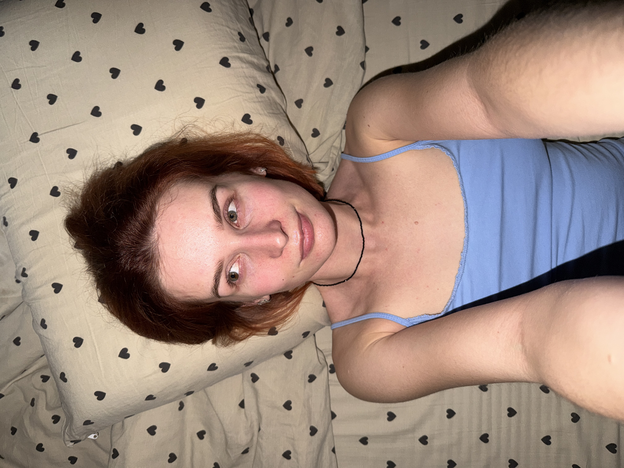 A reddish-haired woman lies on bedding with a heart pattern. She is wearing a blue tank top and a thin black necklace. The scene appears to be indoors. Location information not present.