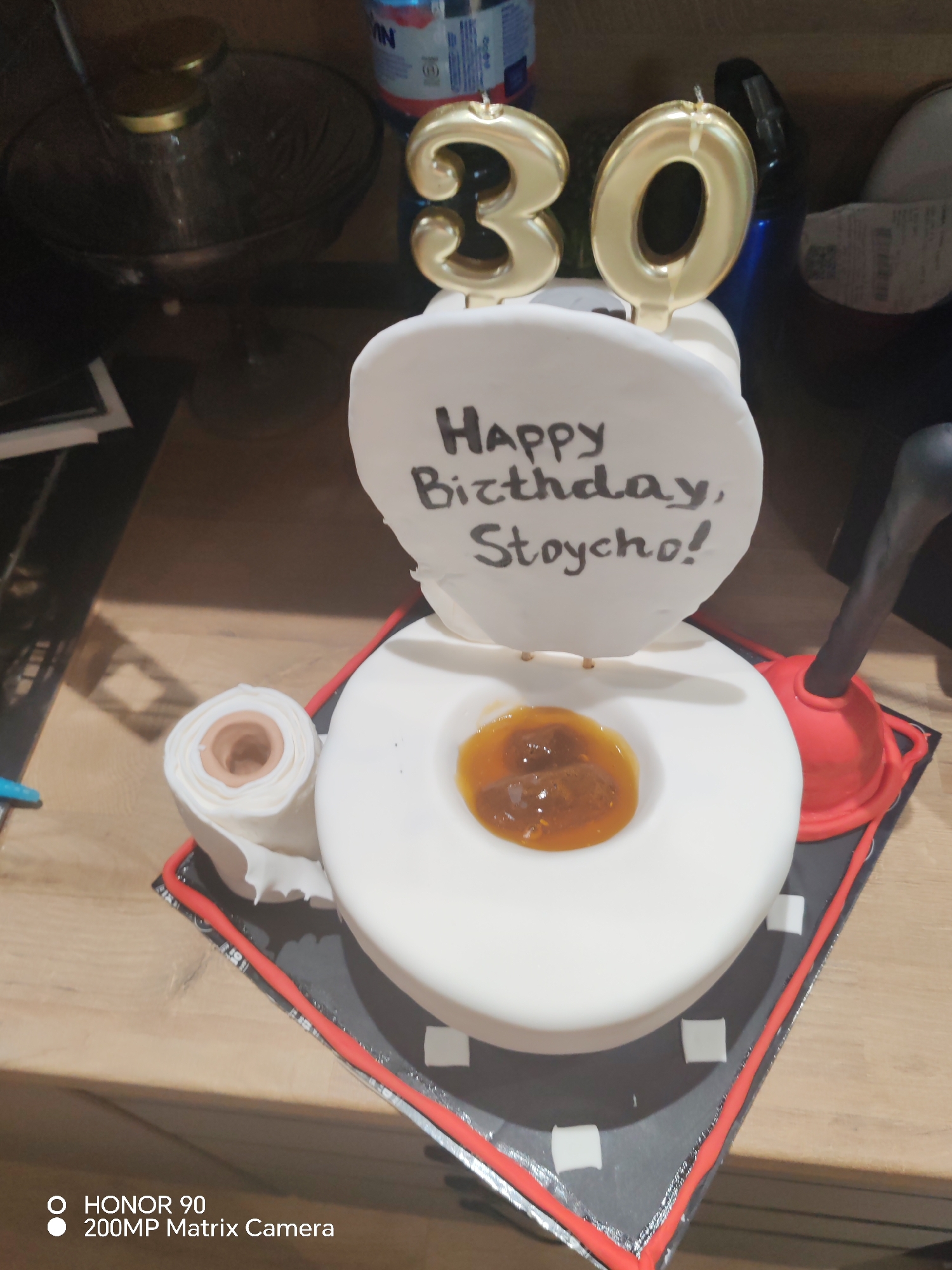 The image captures a humorous novelty cake, meticulously designed to resemble a white toilet, positioned centrally on a cake board. Gold-colored candles forming the number "30" are prominently placed at the back of the toilet cake, indicating a 30th birthday celebration. The toilet seat lid is open, displaying a handwritten message in black script: "Happy Birthday, Stoycho!"
Inside the toilet bowl, there is a dark orange to brown, viscous substance, which appears to be jam or caramel, with some solid inclusions, intended to mimic a crude visual effect. Complementing the toilet theme, a realistic replica of a toilet paper roll, white with a brown core, is situated to the left of the toilet. To the right, a red and black plunger stands upright, further enhancing the playful bathroom motif.
The cake rests on a square cake board featuring a dark grey surface adorned with small white squares, and a thin red border outlining its edge. The background is softly blurred, revealing an indoor setting with a dark glass bowl topped with a golden lid, various bottles (one with red and blue labeling, another blue), and other indistinct items on a light brown wooden surface. The scene suggests a lighthearted birthday event, possibly in a home or party venue in Sofia, Bulgaria, though no specific visual cues for the city are present. A "HONOR 90 200MP Matrix Camera" watermark is visible in the bottom left corner of the image.