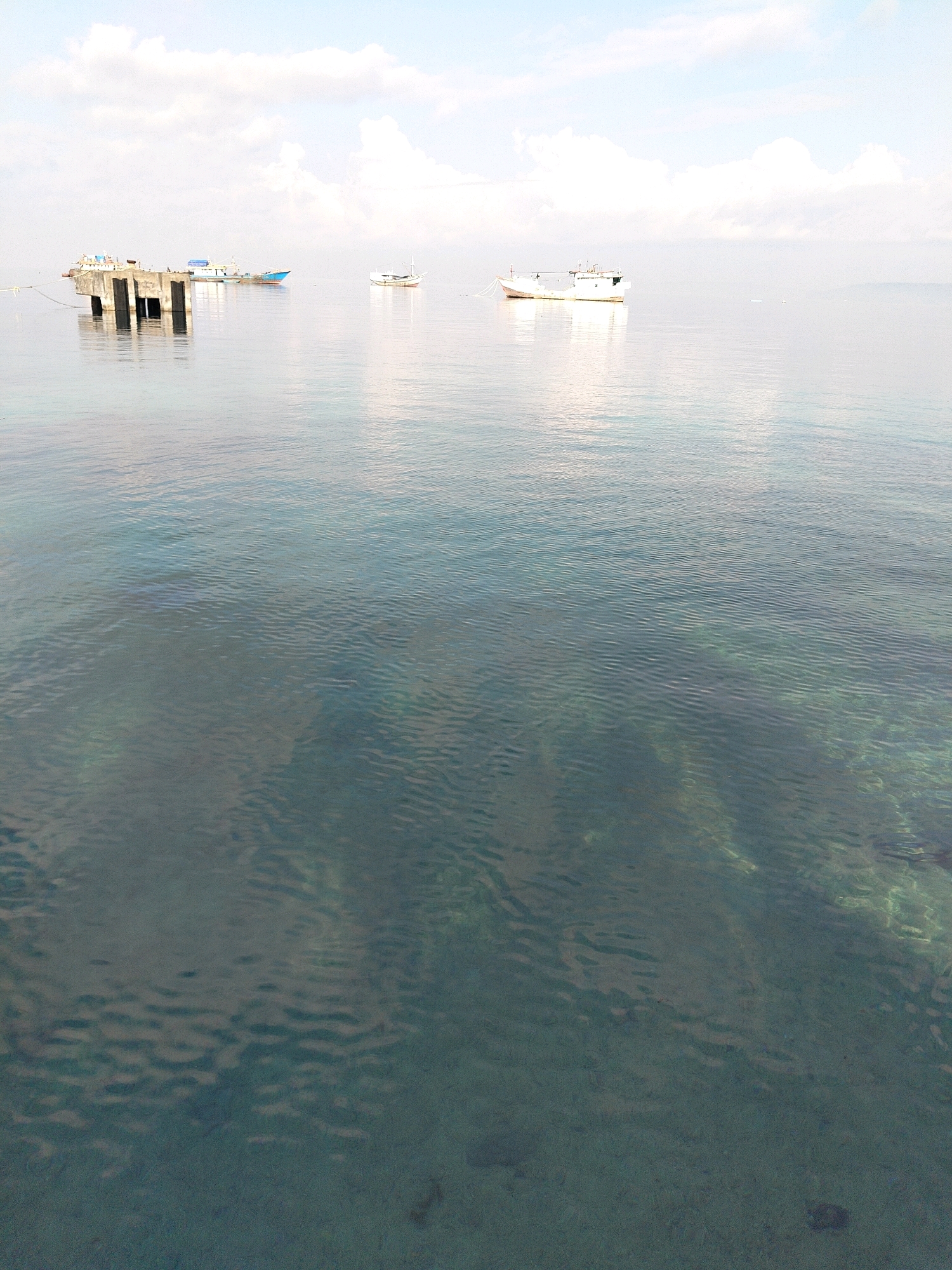A wide shot of the ocean off the coast of Biak, Indonesia, features a clear day with calm waters and scattered clouds in the sky. In the distance, a concrete pier extends into the water, with a few boats moored around it. The water is shallow enough to see the sandy bottom and some coral formations, indicating a tropical location. The overall impression is one of tranquility and natural beauty, with the presence of human structures suggesting a port or a coastal village.