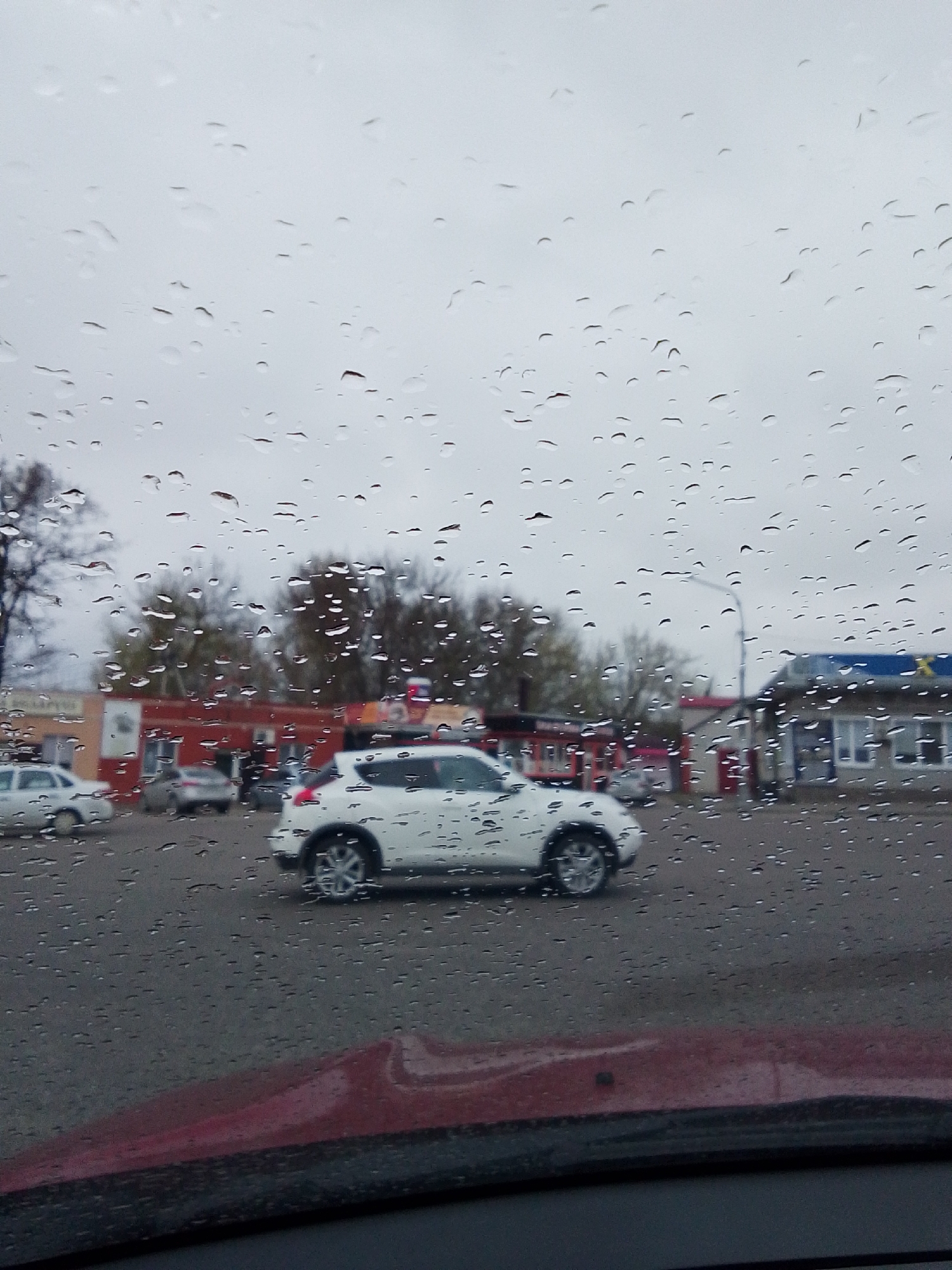 The image shows a street scene in Khlevnoye, Russia, viewed through a rain-streaked windshield. A white Nissan Juke is in motion on the wet asphalt, with a few other vehicles parked or moving in the background. Buildings are visible, including a structure with a blue roof and a large yellow "X" sign on its side, and a red building with red and white signage. Trees are scattered in the background, and the sky is overcast. The dashboard of the car from which the picture is taken is visible at the bottom of the image.