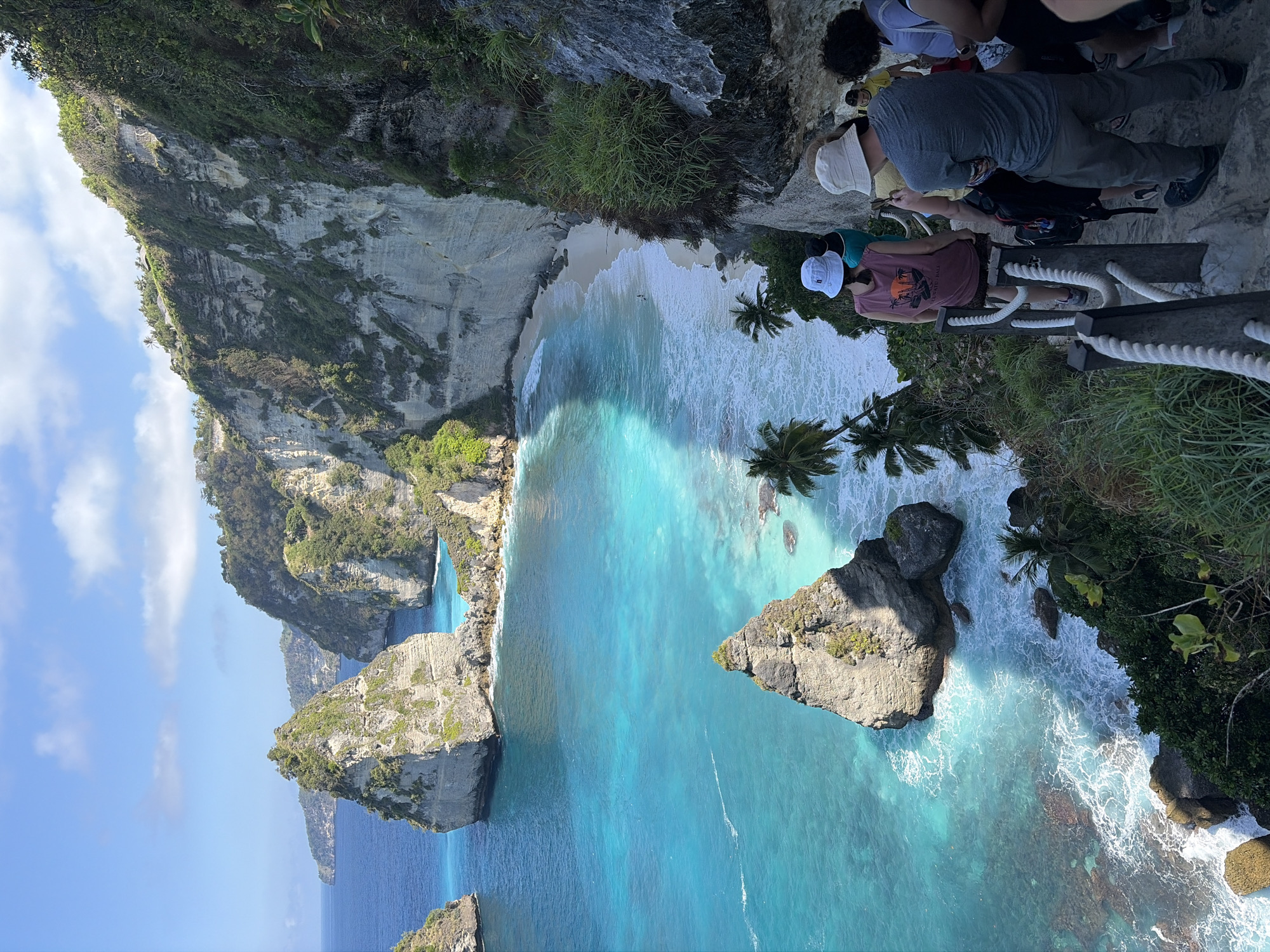 The image shows a scenic coastal view of Nusa Penida, Indonesia, specifically the viewpoint overlooking Diamond Beach. In the foreground, a group of people are walking down a steep, rocky path lined with a rope railing. The path descends towards a pristine white sand beach bordered by turquoise waters. Several large rock formations, known as "diamonds," jut out from the sea in the distance. The cliffs surrounding the beach are covered in lush green vegetation. The sky is blue with scattered white clouds, indicating a sunny day. The water is clear and vibrant, with waves crashing onto the shore. The overall scene is one of natural beauty and popular tourist destination.