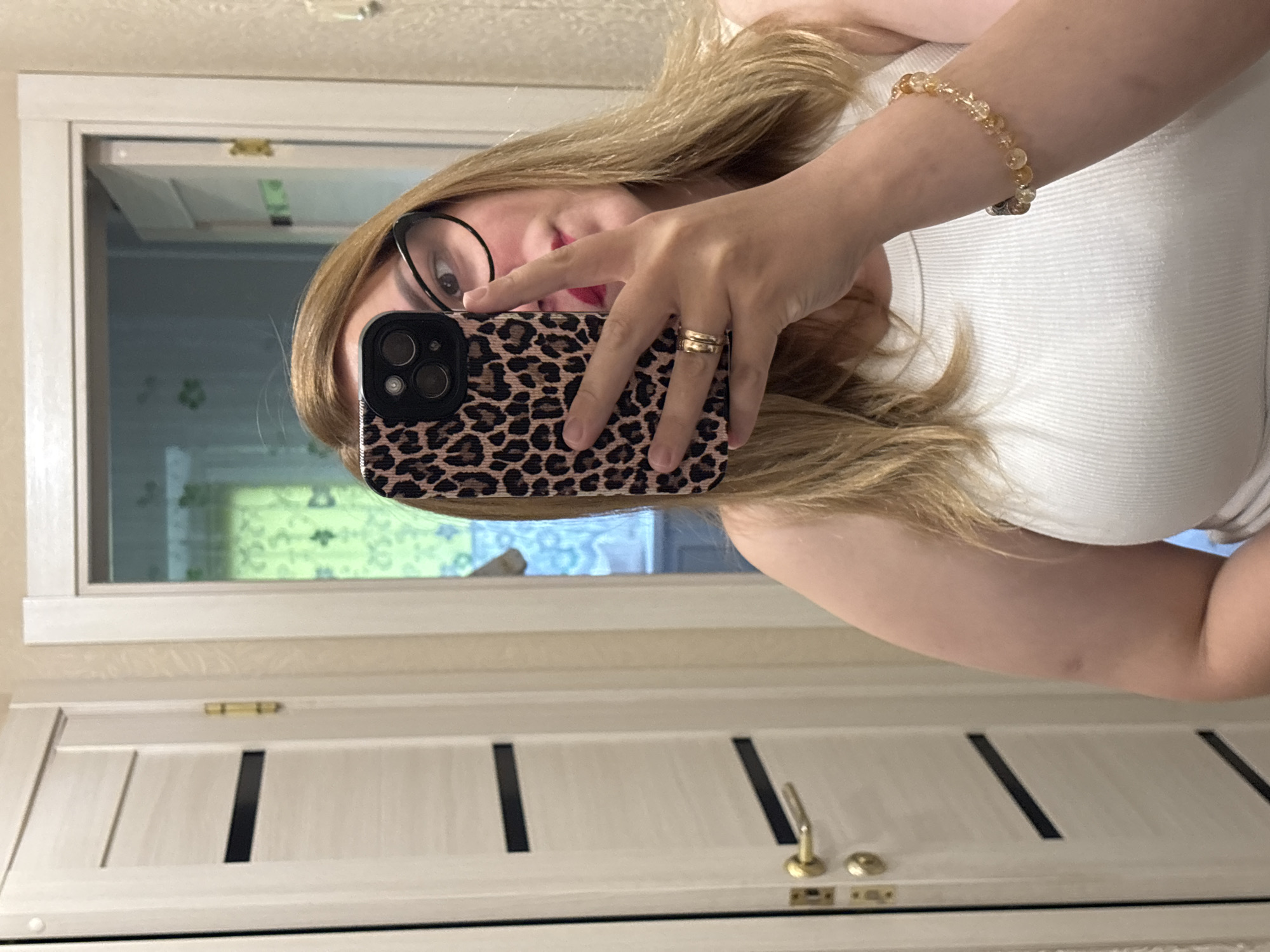 A woman is taking a mirror selfie in what appears to be a home hallway. She has blonde hair and is wearing glasses and a white tank top. She is holding a phone with a leopard print case. On her left hand, she is wearing two gold rings, and on her right wrist, she has a beaded bracelet. To her left is a white door with black stripes and a gold handle. The background through the doorway appears to be a room with green curtains.