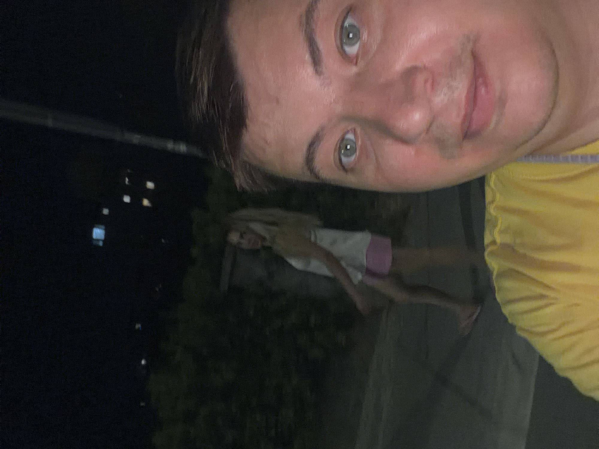 A selfie taken at night on a paved walkway in Budva, Montenegro, features a man in the foreground wearing a yellow shirt. Behind him, a woman with blonde hair is walking away from the camera, wearing a white top and pink shorts. In the background, blurred lights indicate distant buildings. The scene is dimly lit, with the overall impression being that of an outdoor, urban setting after dark.
