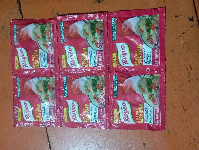 Six red packets of Royco Bumbu Kaldu Rasa Ayam (Chicken Broth Seasoning) are neatly arranged in two rows of three on a textured, brownish-orange surface, likely a wooden floor or table in Pubian, Indonesia. Each rectangular packet features the white "Royco" logo and the text "SINCE 1973." An illustration of a cooked chicken drumstick in a bowl of broth with green vegetables is prominent on the front.
Each packet displays the product name "BUMBU KALDU RASA AYAM" and indicates a "FORMULA BARU!" (New Formula!). The price "Rp500" (Indonesian Rupiah) is visible on a yellow banner. A "HALAL" certification logo, with "INDONESIA" below it on some packets, is positioned on the right side. Smaller text on the packaging includes phrases like "Komitmen Kami" (Our Commitment) and descriptions of the flavor, such as "Rasa Ayam yang Nikmat" (Delicious Chicken Flavor) and "Aroma Ayam Kuat & Enak" (Strong & Delicious Chicken Aroma). The packets appear new and unopened, presented as a straightforward product display.