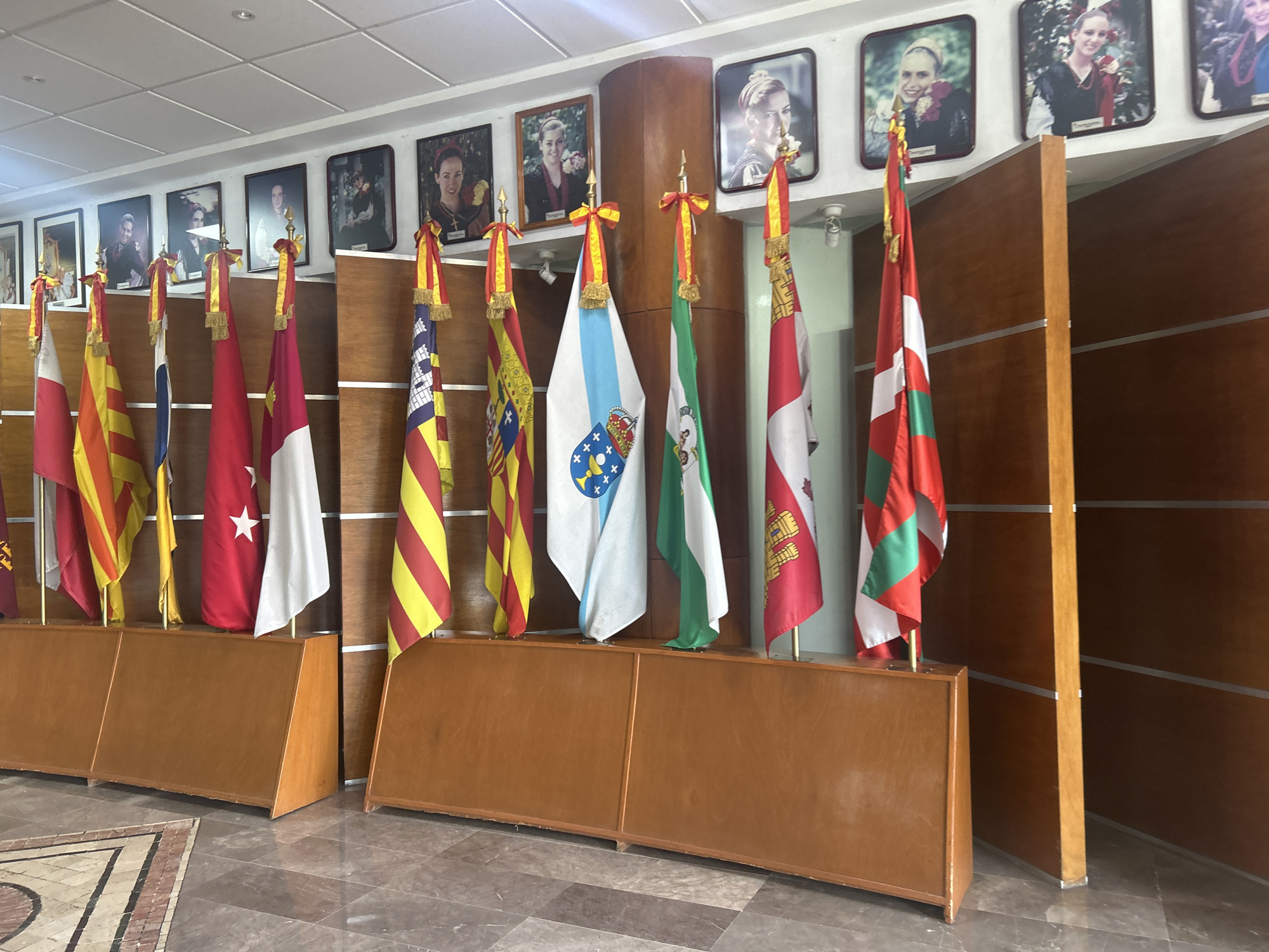 The image shows a collection of flags on display within a building. There are ten flags in total, each mounted on a wooden base. The flags are arranged in a row, and they are hung from decorative golden poles with red and gold ribbons. Behind the flags, there is a wall decorated with framed portraits. The floor is tiled with a gray and beige patterned design. The lighting in the room is bright.
The building is likely located in the city of Puebla, Mexico. This is supported by the flags that are present in the photograph. These flags include the flags for the city of Puebla, Catalonia, and other regions and countries.